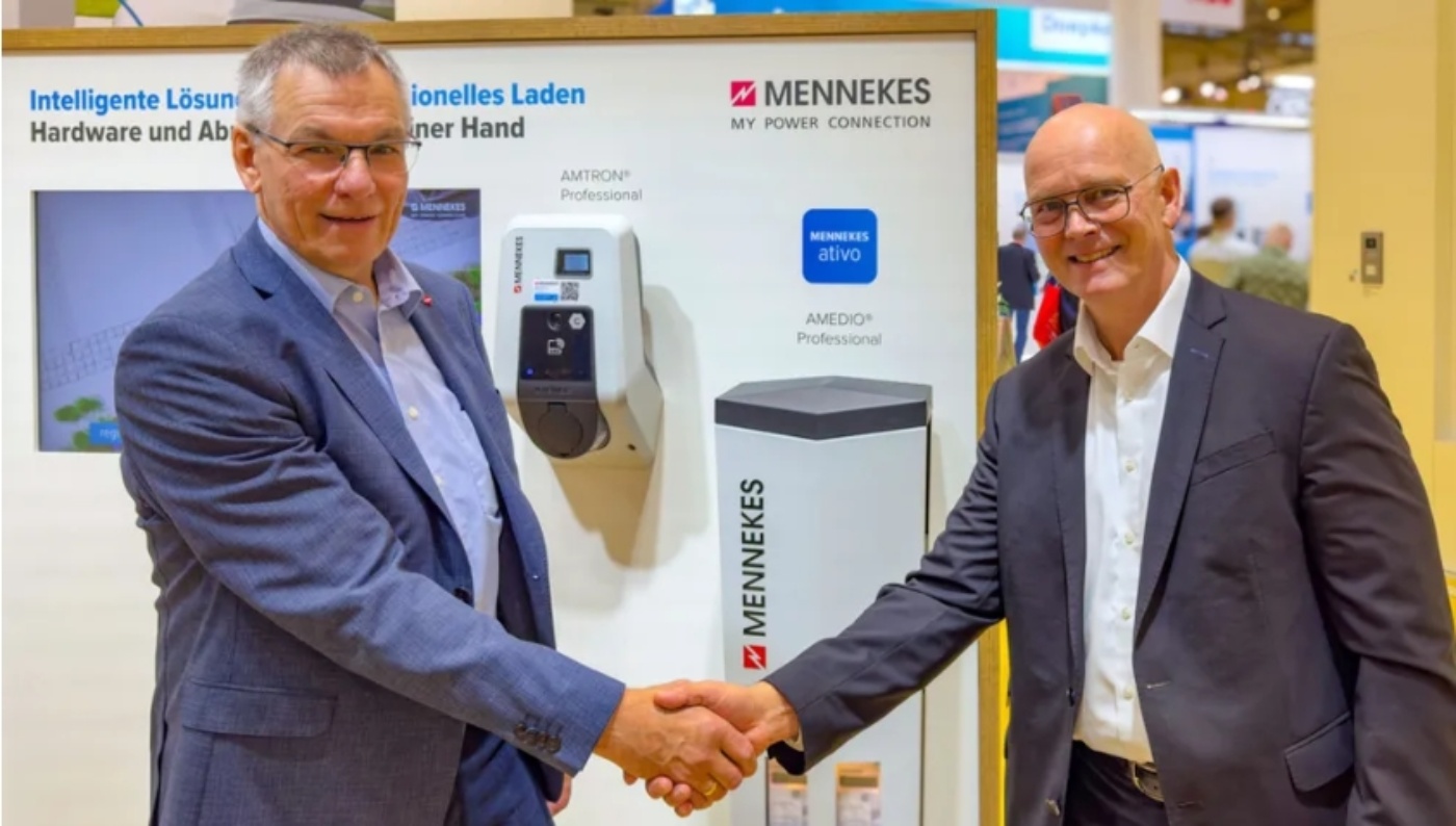 Two men in business attire shake hands in front of a display for Mennekes electric vehicle charging solutions at a trade fair.