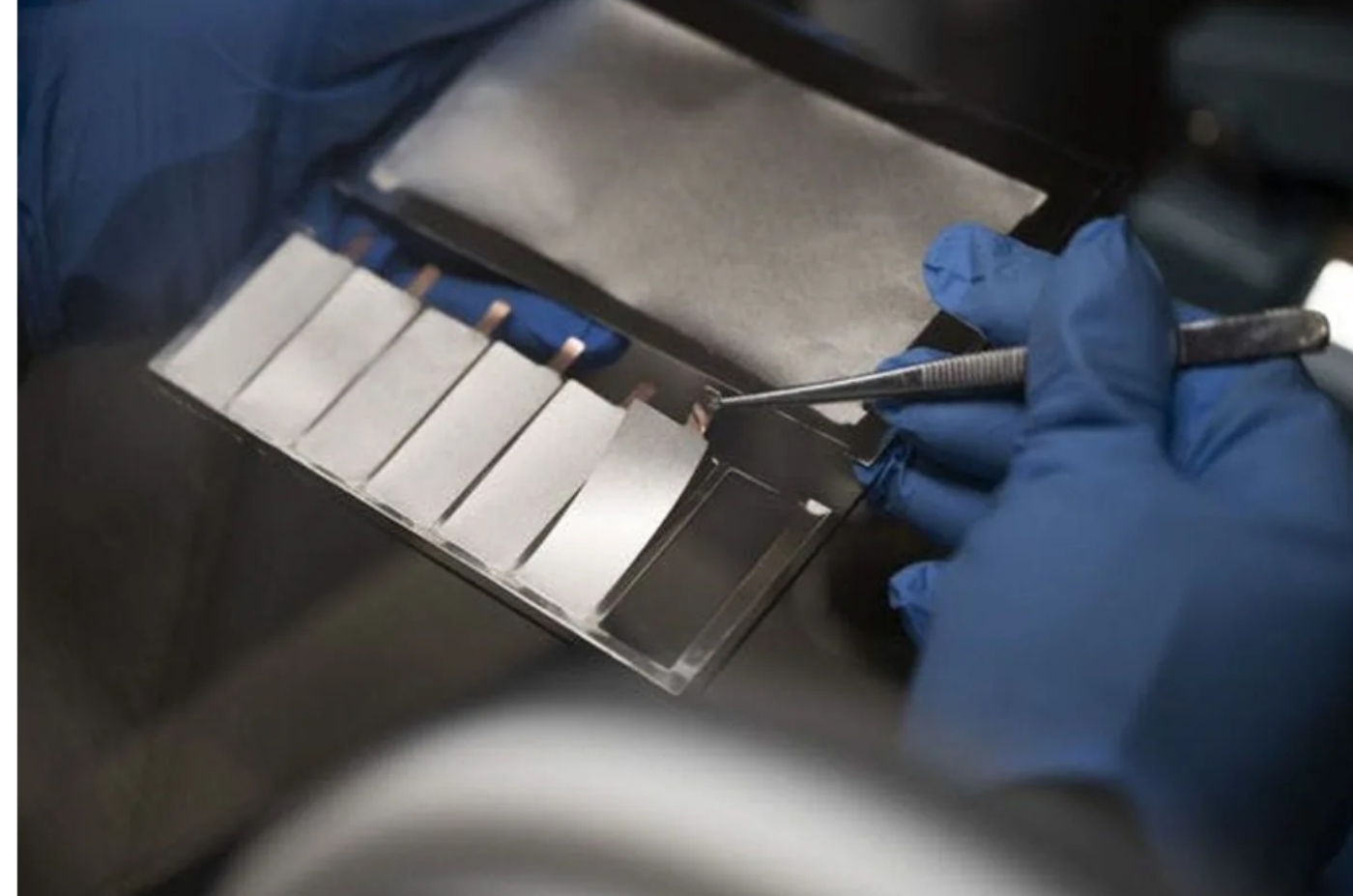 Gloved hands handle a lab sample of battery cell components using tweezers inside a controlled environment.