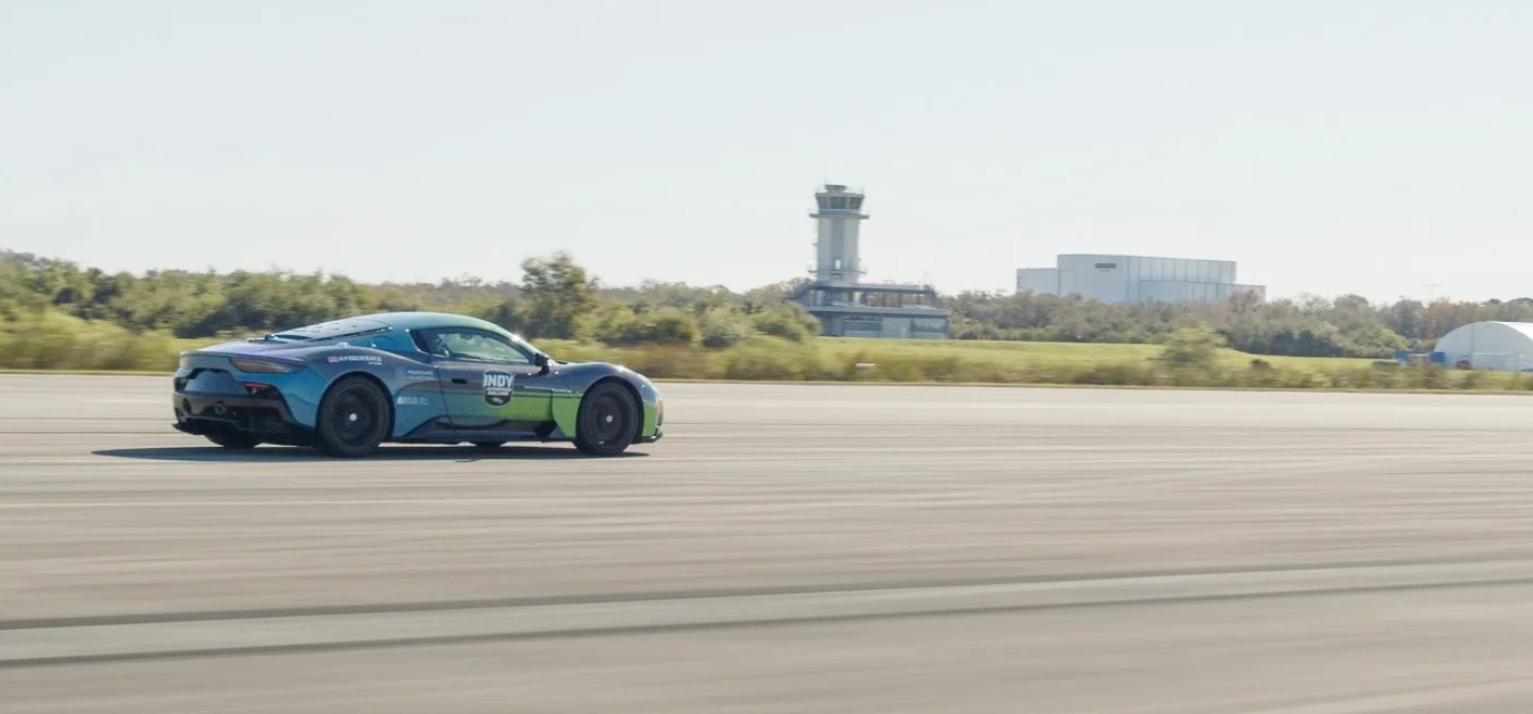 Side view of a high-performance Maserati sports car driving on an airstrip, with control tower and hangars in the background.