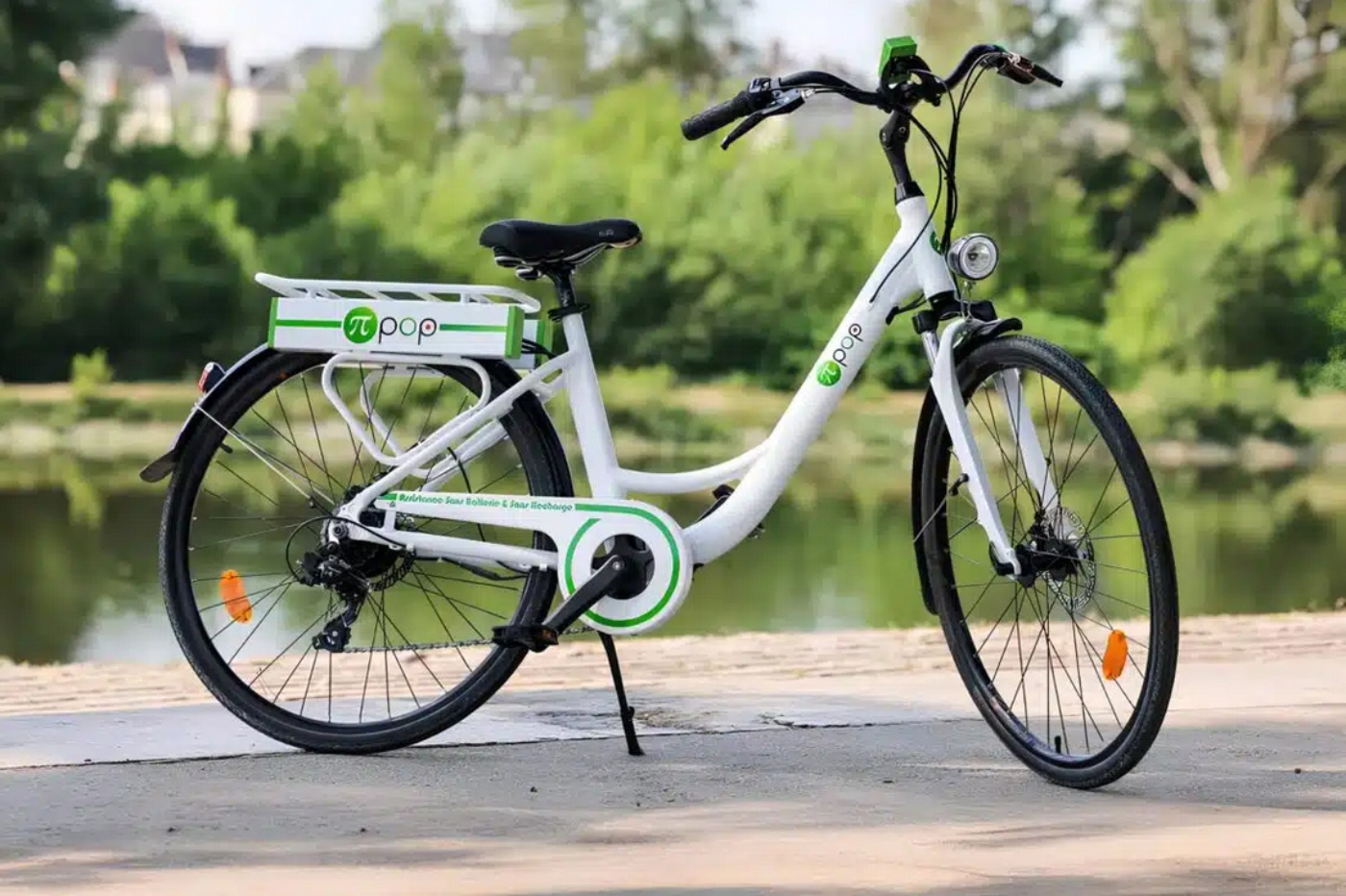 A white Pi-Pop electric bicycle with green accents is parked on a path near a lake, featuring a rear battery pack and front headlight.