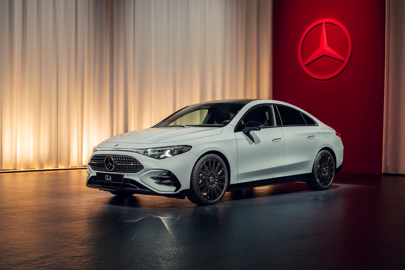 A white Mercedes-Benz CLA sedan is displayed in a showroom with soft lighting and a large red wall featuring the Mercedes-Benz logo.