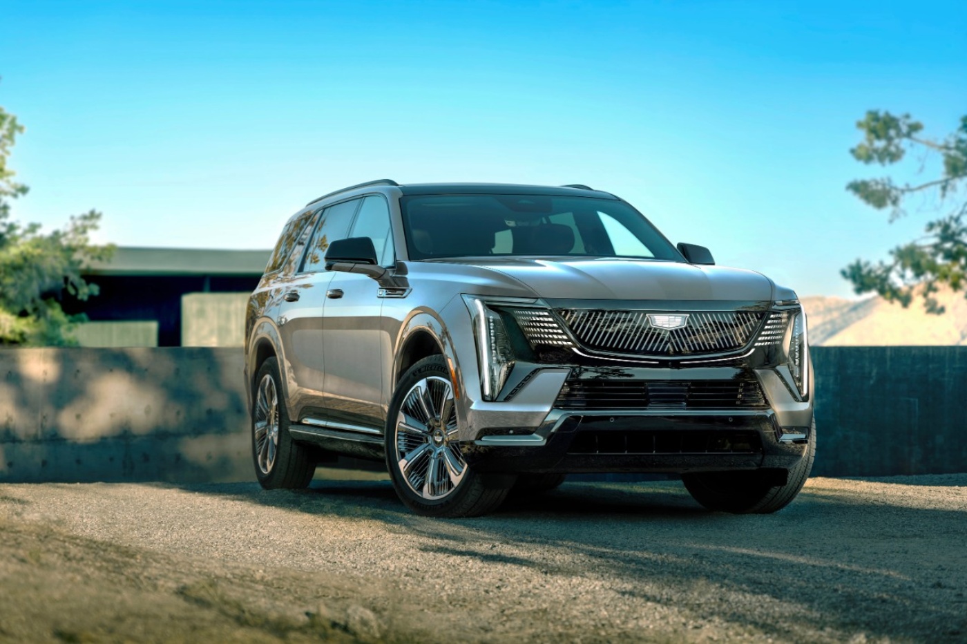 A silver Cadillac electric SUV is parked outdoors in front of a low wall, shown in bright daylight with trees and hills in the background.