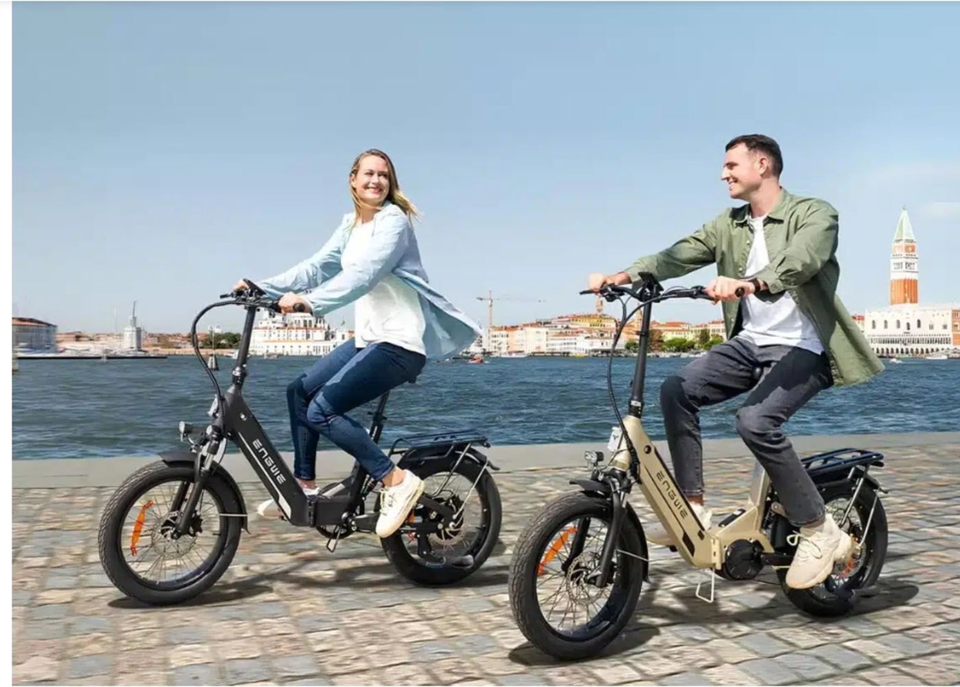 Eine Frau und ein Mann fahren auf klappbaren E-Bikes an einer Uferpromenade entlang, im Hintergrund eine historische Stadt mit Glockenturm.