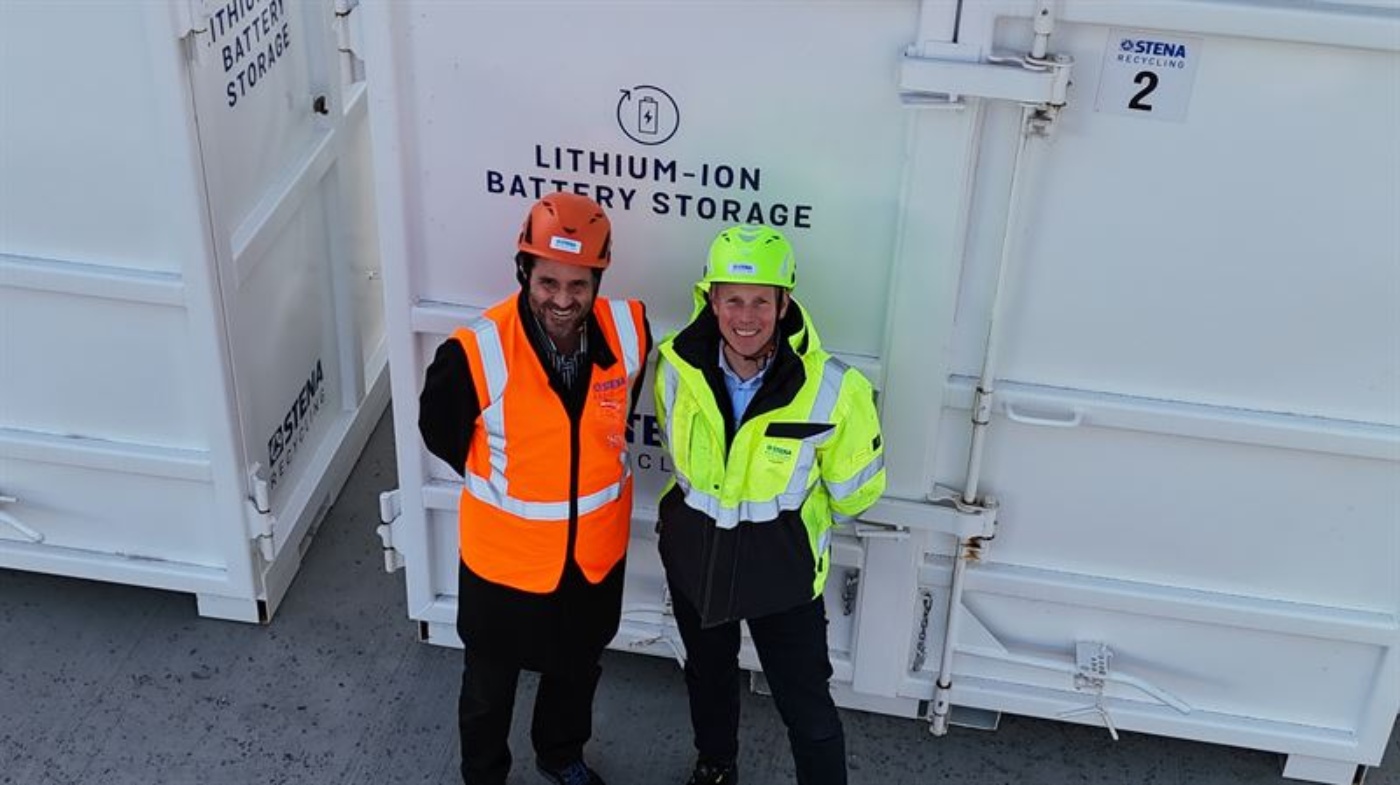 Zwei Personen in Sicherheitskleidung stehen vor einem Container mit der Aufschrift „Lithium-Ionen-Batteriespeicher“ in einer Recyclinganlage.