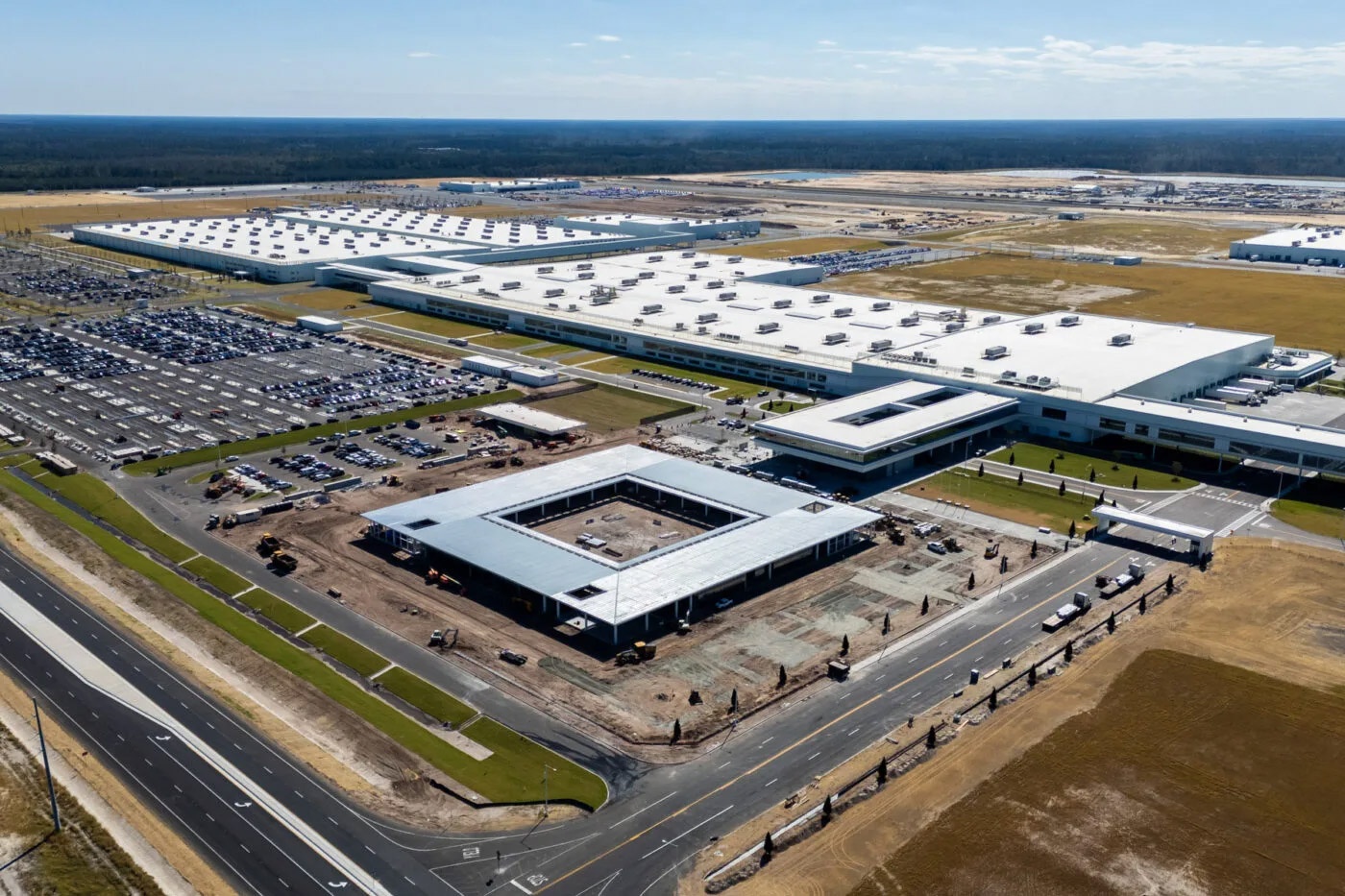 Aerial view of a large industrial complex with white-roofed factory buildings, parking areas, and construction zones, surrounded by open land.