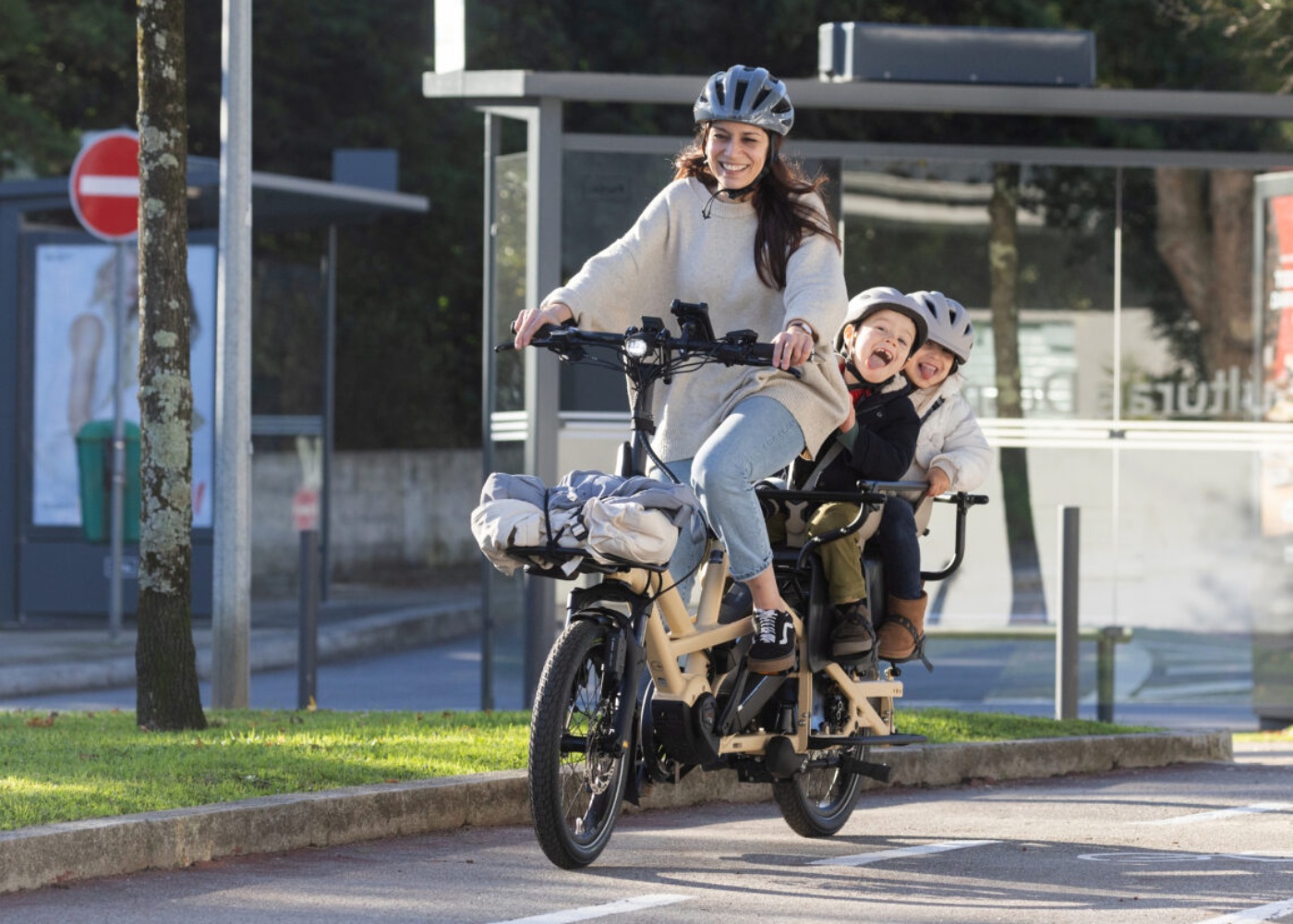 A woman rides a beige electric cargo bike with two children seated behind her, all wearing helmets, on a sunny urban street.
