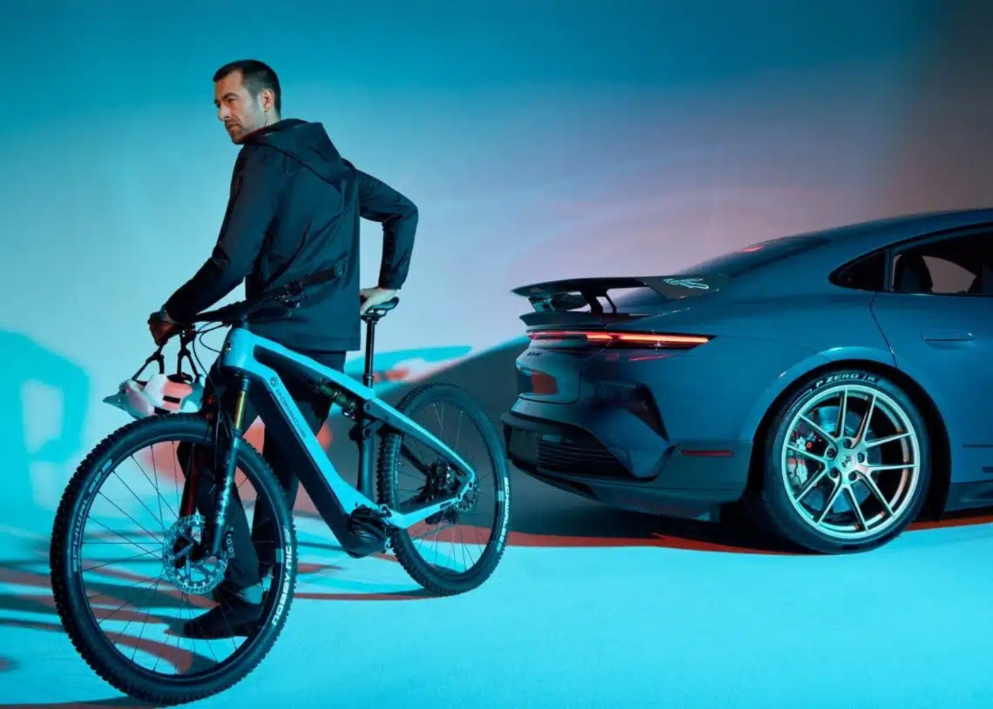 Man with a light blue electric mountain bike stands next to a blue Porsche sports car in a studio with dramatic lighting.