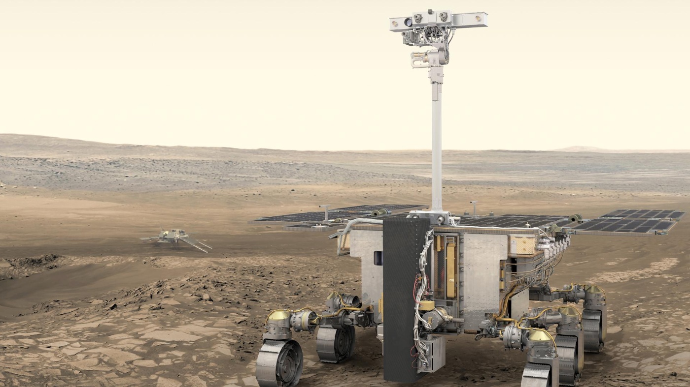 Ein sechsrädriger Marsrover mit ausgeklappten Solarpanels steht auf einer felsigen Marslandschaft, im Hintergrund ist eine Landesonde zu sehen.