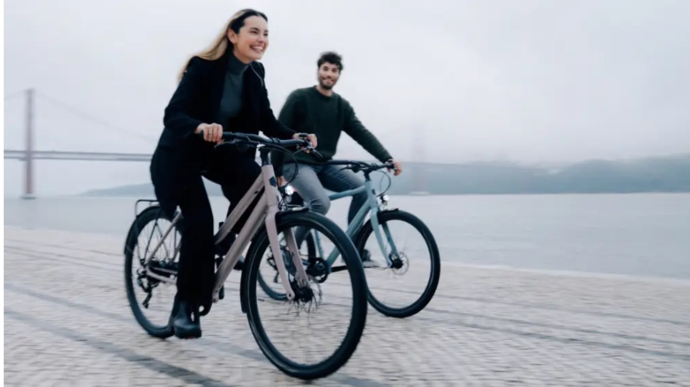 Zwei Personen fahren bei bewölktem Wetter lachend auf E-Bikes an einer Uferpromenade entlang.