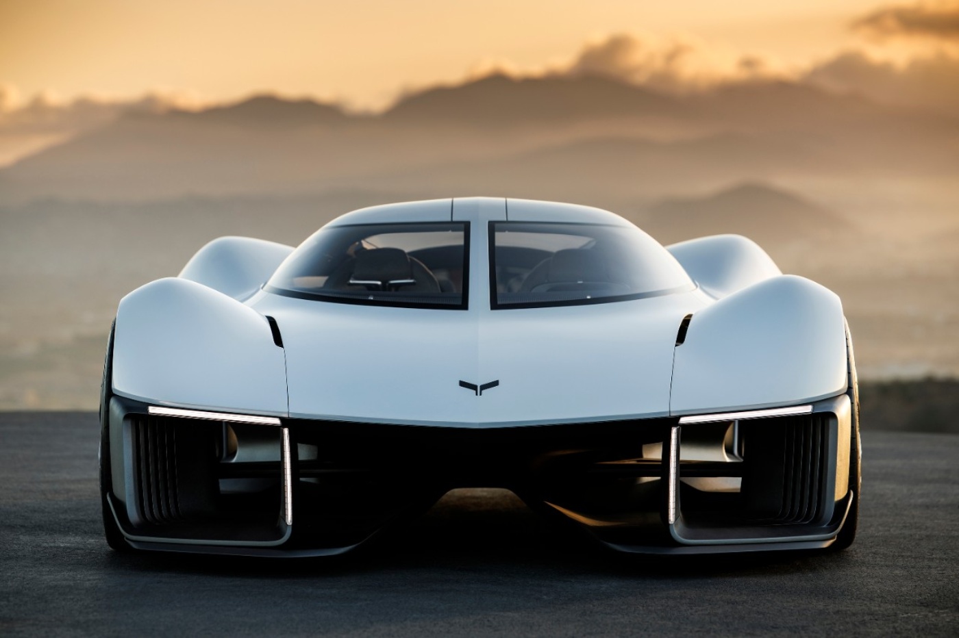 Futuristic white electric sports car seen from the front, parked on a road at sunset with mountains in the background.