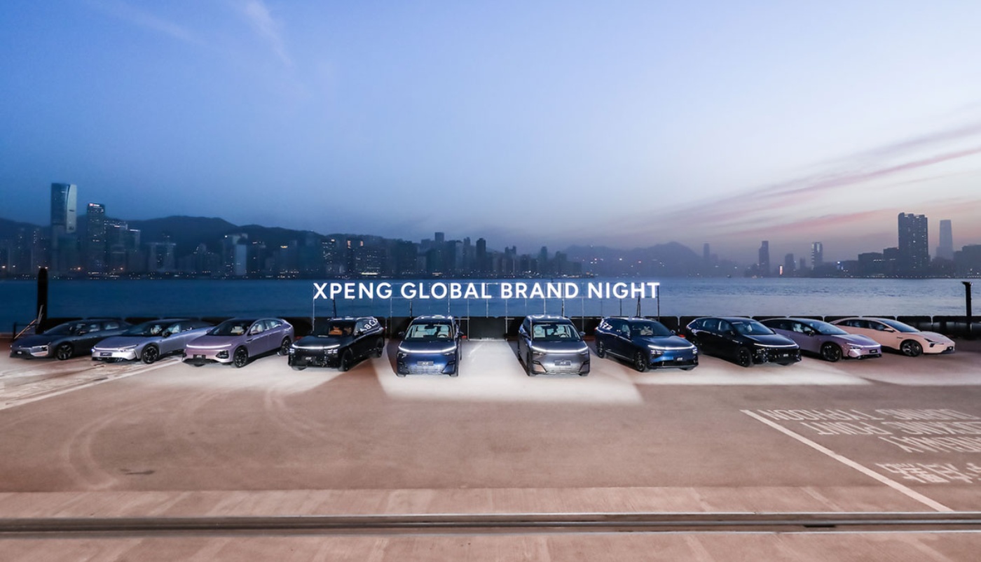 Line-up of electric cars at the XPeng Global Brand Night event, with a city skyline and waterfront in the background.