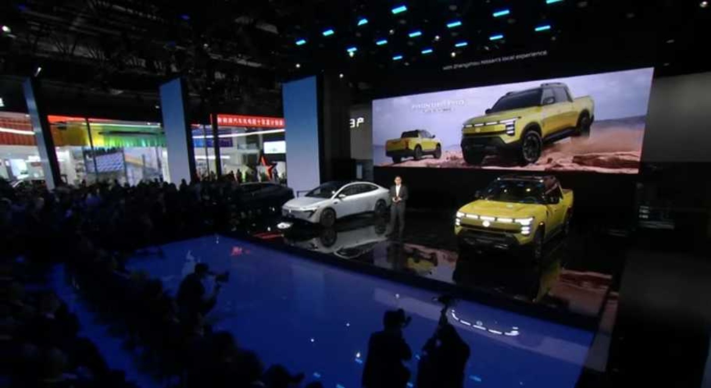 Eine Ausstellungsbühne mit drei Elektrofahrzeugen, darunter ein gelber SUV im Vordergrund, ein weißer Crossover und ein großer Bildschirm, der den SUV bei einer Offroad-Fahrt zeigt.