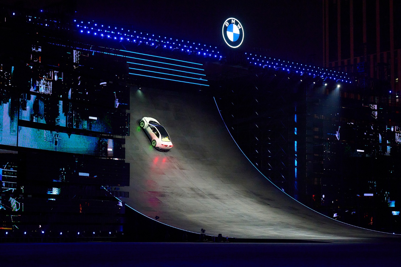 Ein weißes BMW-Fahrzeug mit leuchtenden Felgen fährt bei einer nächtlichen Show eine steil beleuchtete Rampe hinauf, über der das BMW-Logo leuchtet.