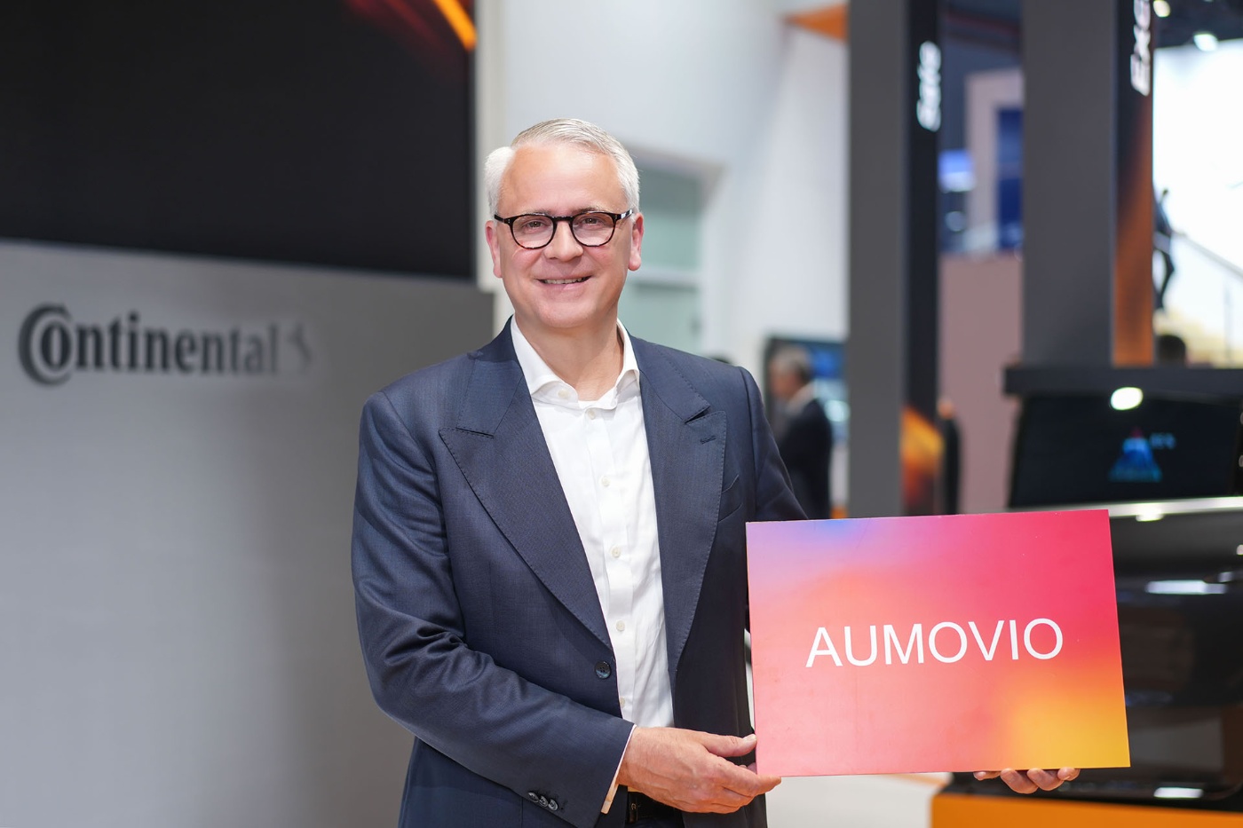 Ein Mann im Anzug steht an einem Continental-Messestand und hält ein farbiges Schild mit der Aufschrift „AUMOVIO“.