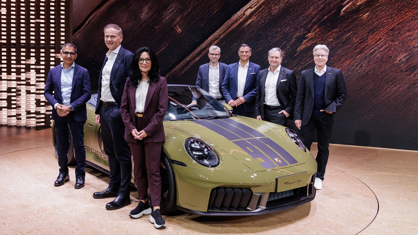 Sieben Personen in Businesskleidung posieren vor einem olivgrünen Porsche-Sportwagen auf einem Ausstellungsboden.
