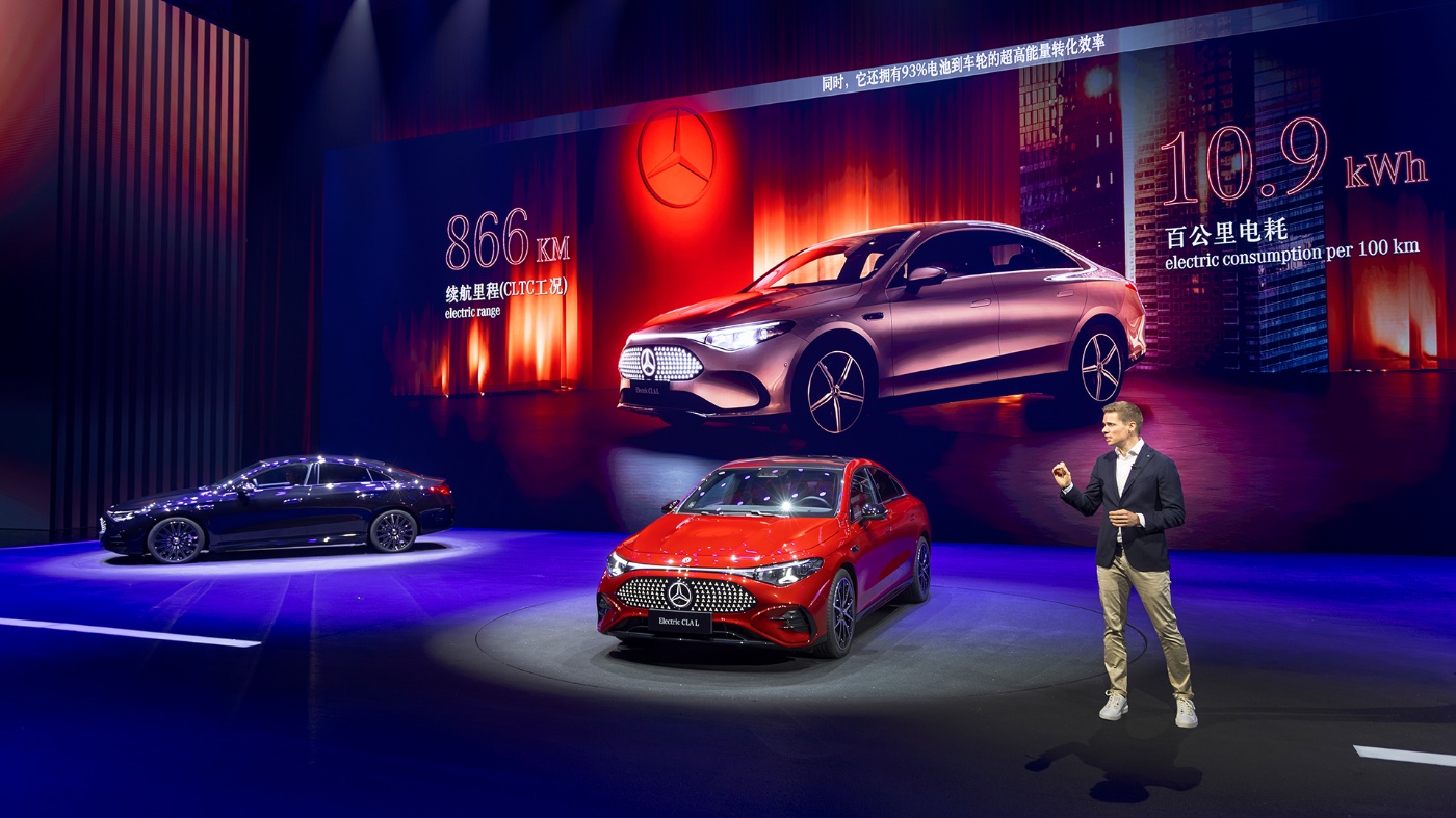 Ein Moderator steht neben einem roten Mercedes-Benz Elektroauto auf einer Bühne, im Hintergrund zeigt ein großer Bildschirm Leistungsdaten wie 866 km Reichweite und 10,9 kWh Verbrauch pro 100 km.