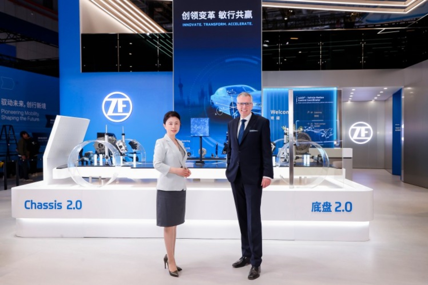 Zwei Personen stehen vor einem modernen Messestand von ZF mit Automobilkomponenten und dem Slogan „Innovate. Transform. Accelerate.“ auf einem großen Bildschirm.