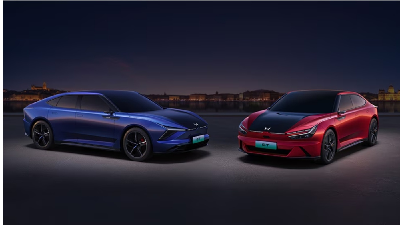 Zwei moderne Elektro-Sportwagen, einer blau und einer rot mit schwarzen Akzenten, stehen nebeneinander bei Nacht vor einer Stadtkulisse.