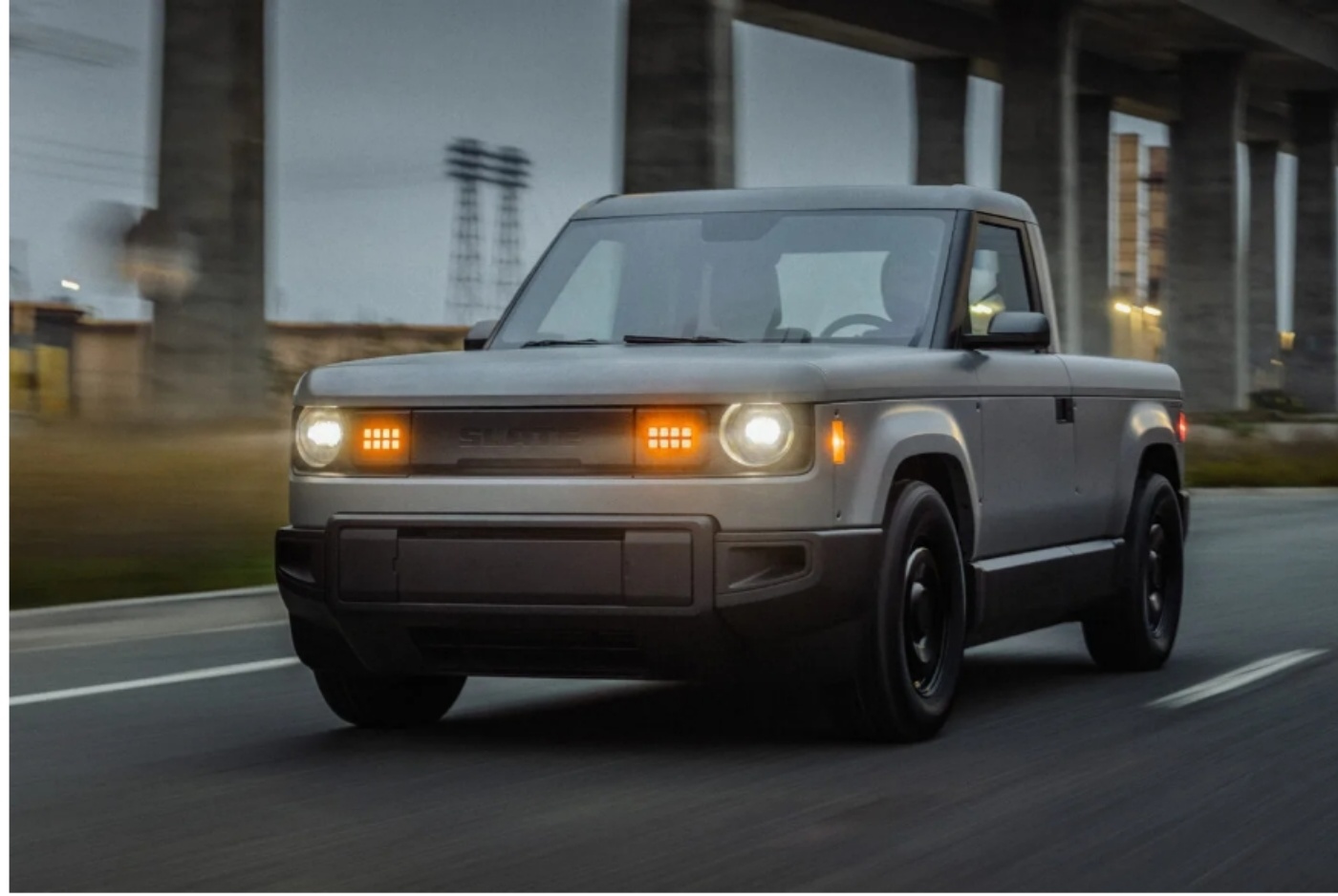 Ein kompakter grauer Elektro-Pick-up mit quadratischen LED-Scheinwerfern fährt bei Dämmerung unter einer Betonbrücke auf einer Straße.