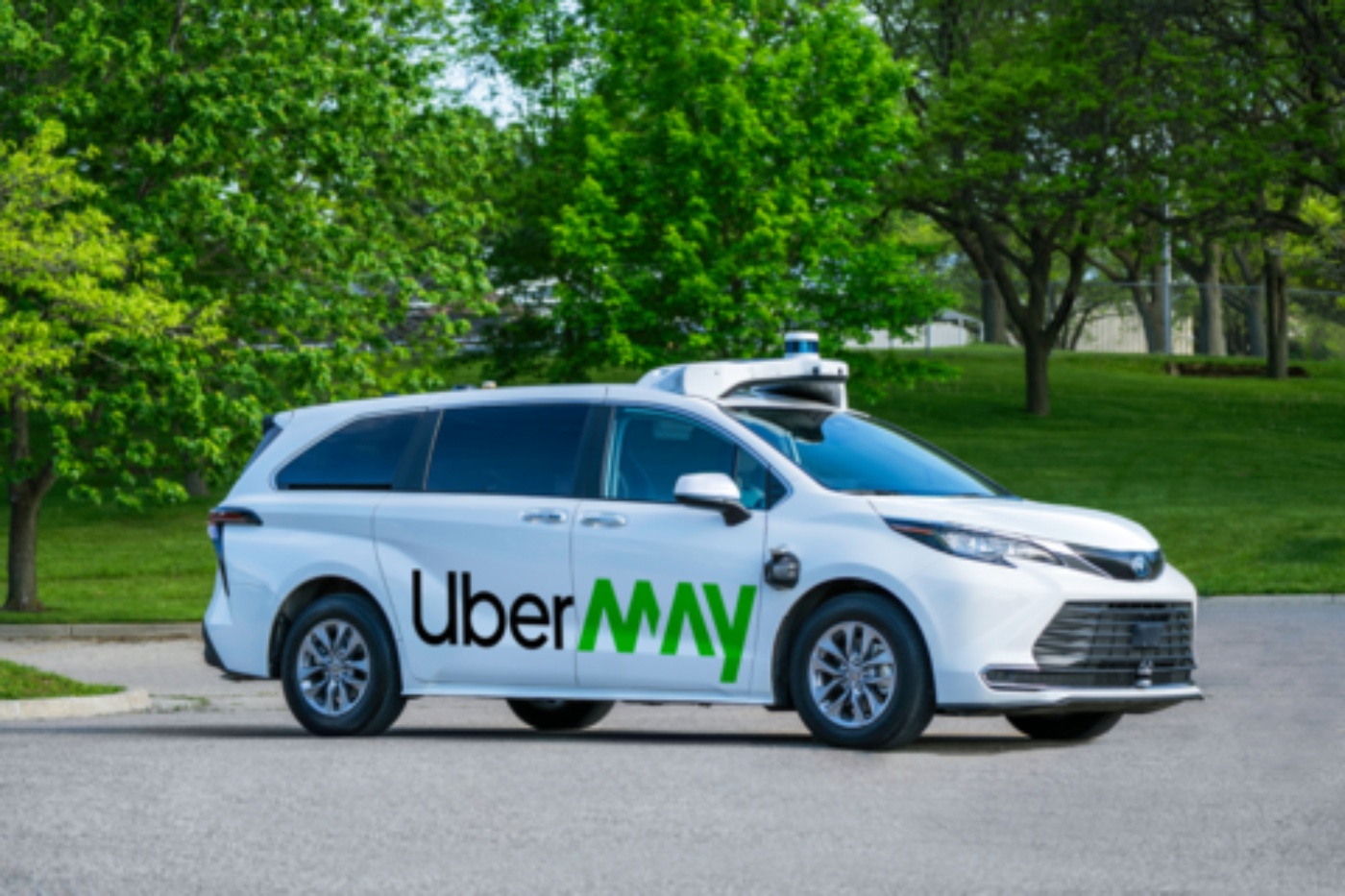 Ein weißer autonomer Minivan mit dem Logo „Uber MAY“ steht auf einer Straße neben einer grünen Parkanlage mit Bäumen im Hintergrund.