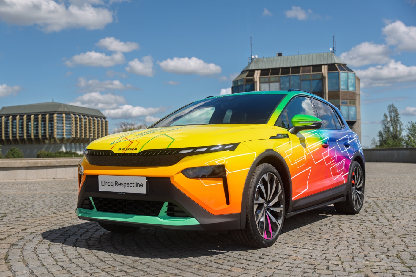 Ein Škoda Elroq Respectline Elektro-SUV steht auf Kopfsteinpflaster und zeigt ein auffälliges Regenbogenfarbdesign mit geometrischen Mustern, bei blauem Himmel und modernen Gebäuden im Hintergrund.