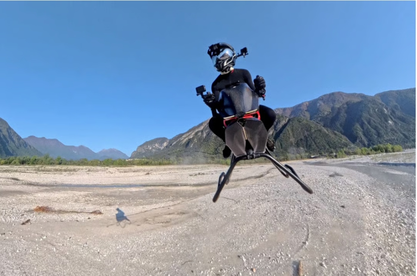 Eine Person in schwarzer Schutzkleidung fliegt mit einem kompakten Hoverbike über ein steiniges, trockenes Flussbett, im Hintergrund sind Berge zu sehen.