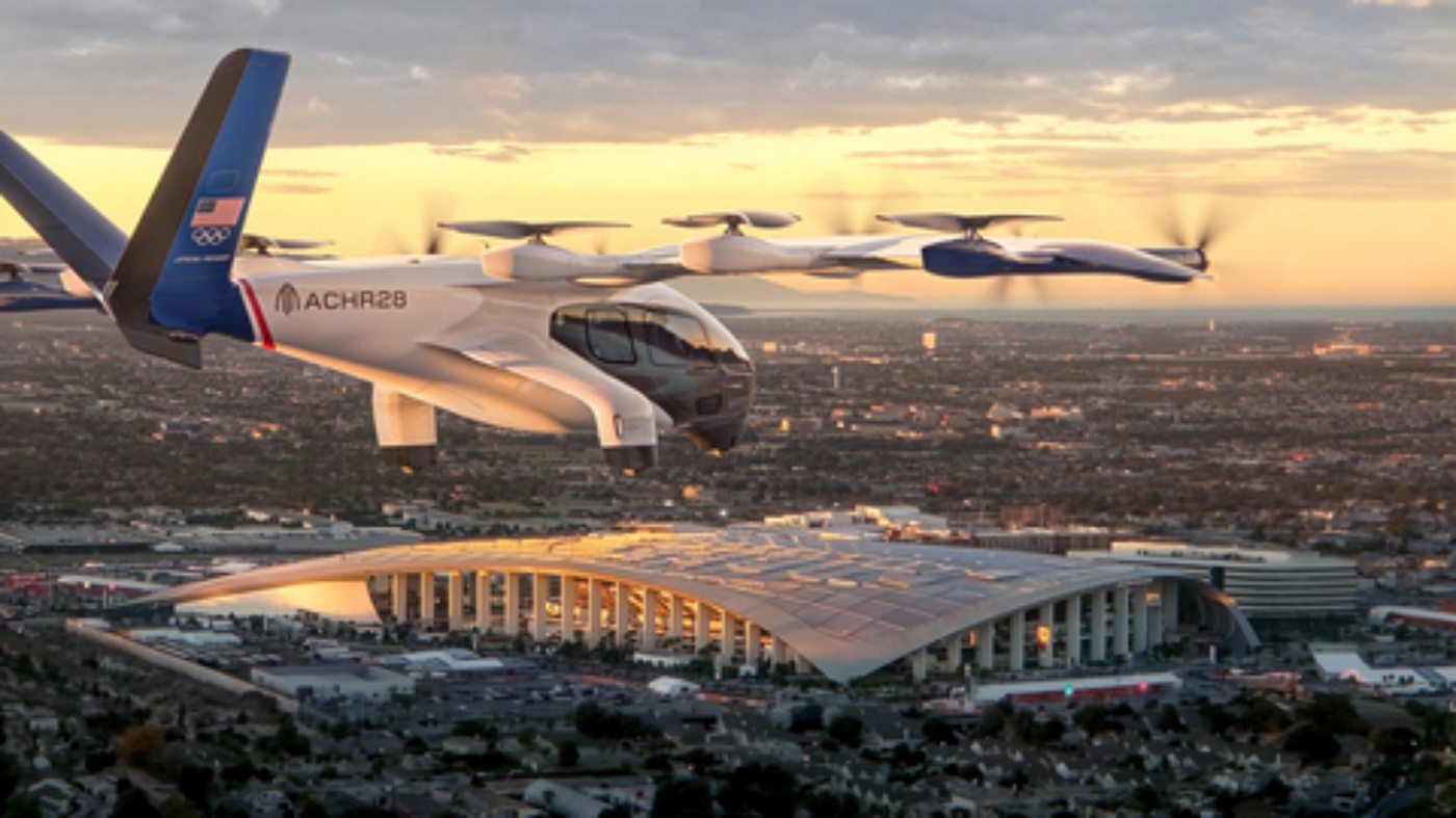 Ein senkrecht startendes Elektroflugzeug fliegt bei Sonnenuntergang über ein großes, modernes Stadion, im Hintergrund erstreckt sich eine Stadtlandschaft.