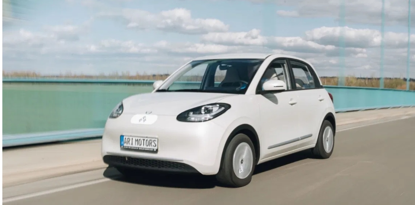 Ein kleiner weißer Elektro-PKW von ARI Motors fährt auf einer Straße mit blauer Leitplanke und bewölktem Himmel im Hintergrund.