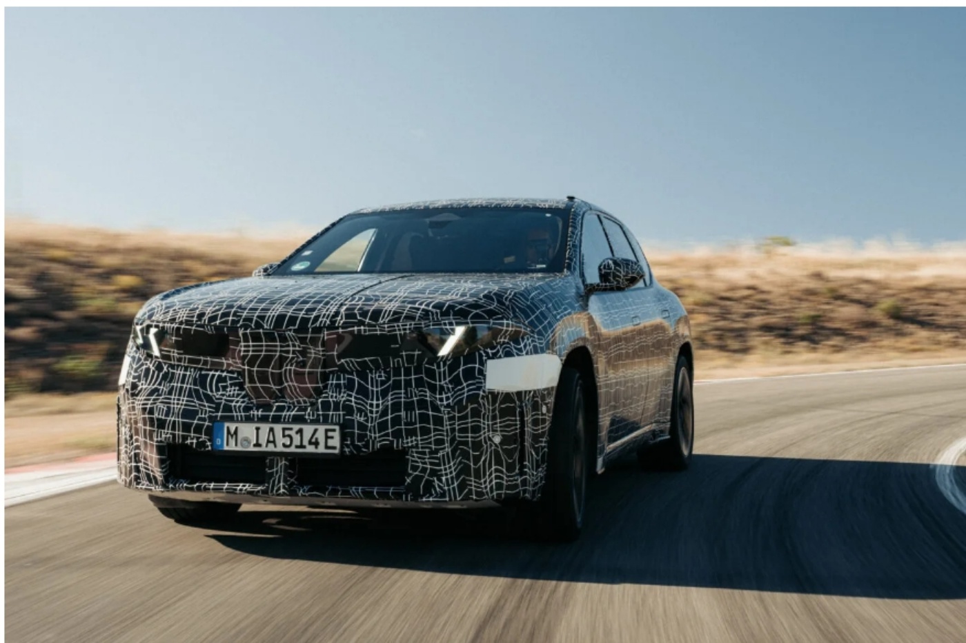 A camouflaged test car drives on a curved road during daytime.