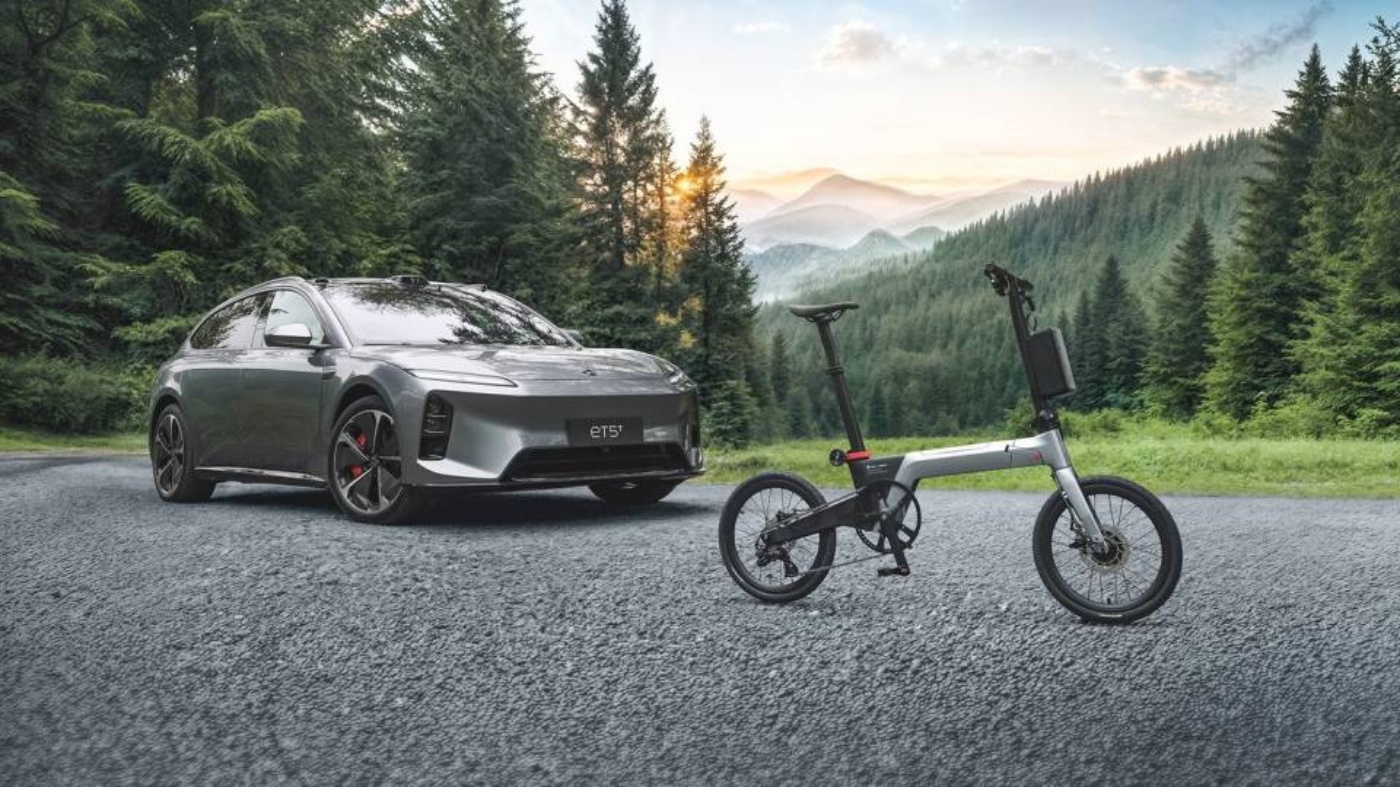 An electric car and a folding e-bike are parked on a road in a forested mountain area.