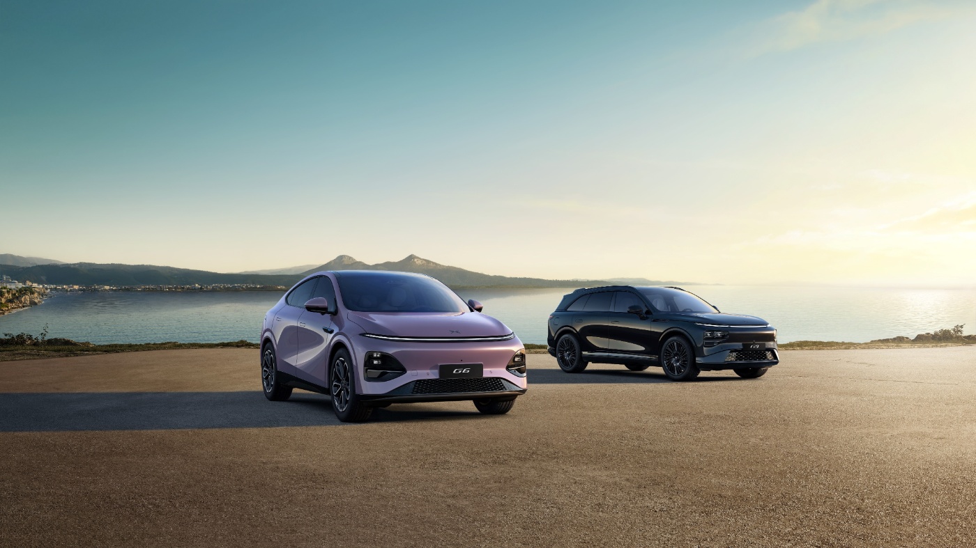 Zwei Elektro-SUVs stehen bei Sonnenuntergang am Meer, mit Bergen und Küstenstadt im Hintergrund.