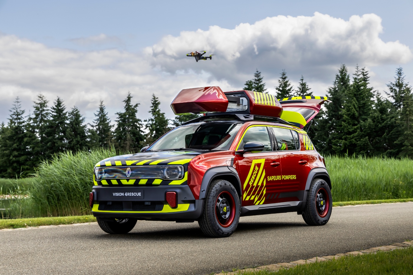 Buntes Renault-Rettungsfahrzeug mit offenem Dachkasten und fliegender Drohne an Waldstraße.