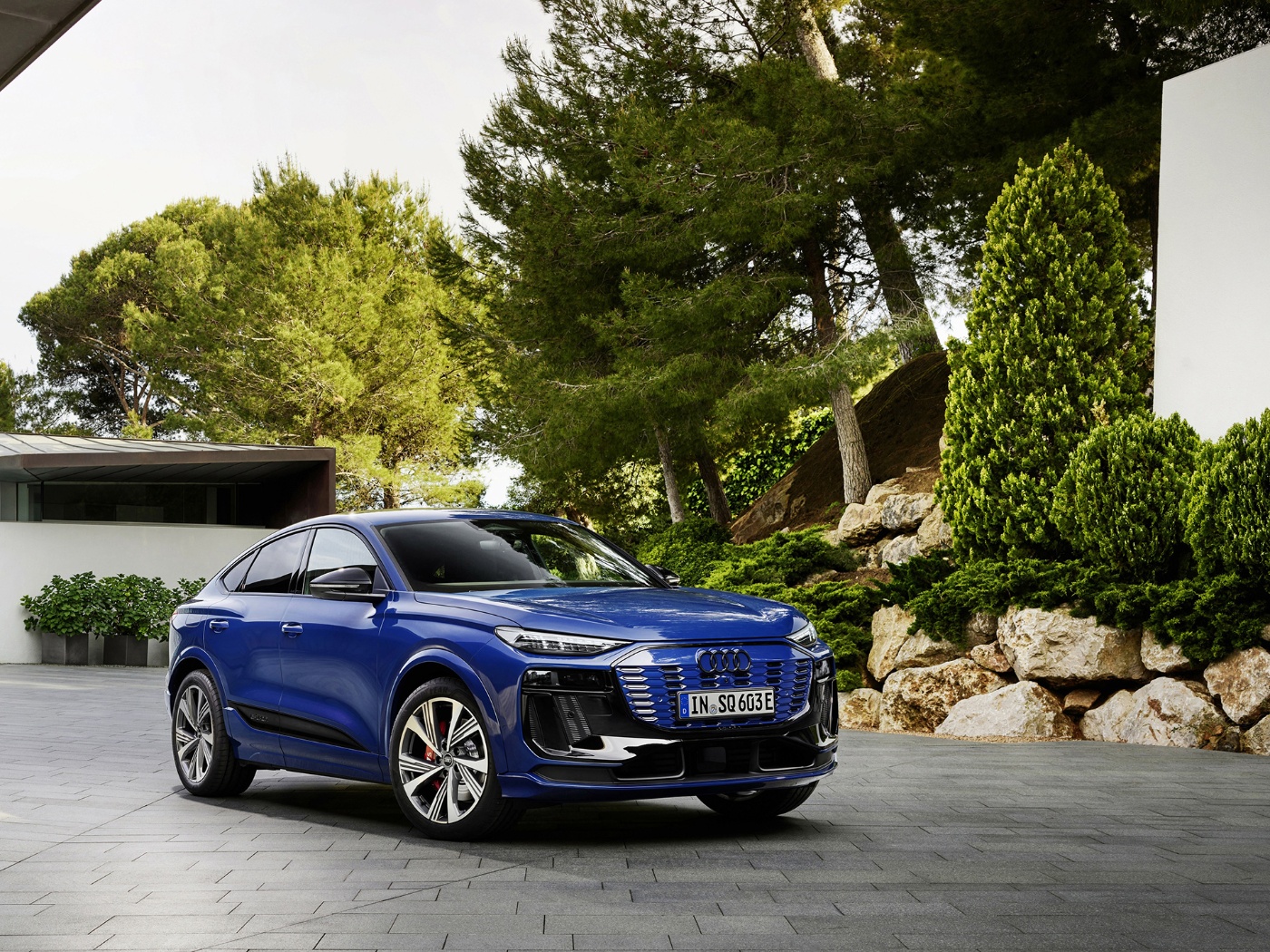 Ein blauer Elektro-SUV steht vor einem modernen Haus mit Bäumen und viel Grün im Hintergrund.