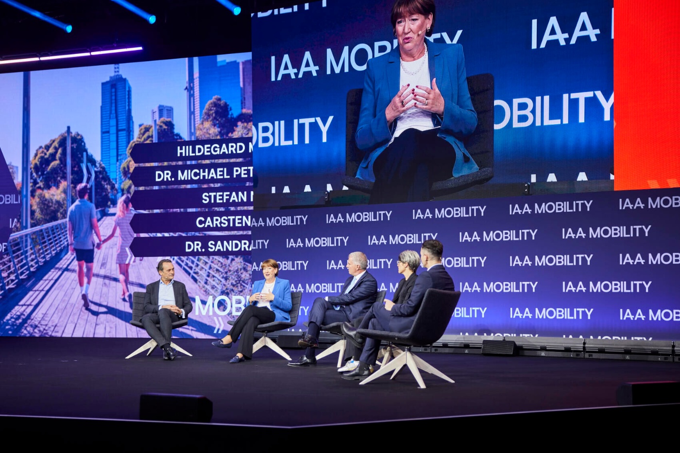 Fünf Personen diskutieren auf der Bühne über Mobilität bei der IAA, eine Person ist groß eingeblendet.