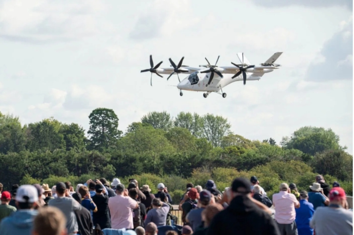 Crowd watching a white electric aircraft with multiple rotors flying low.