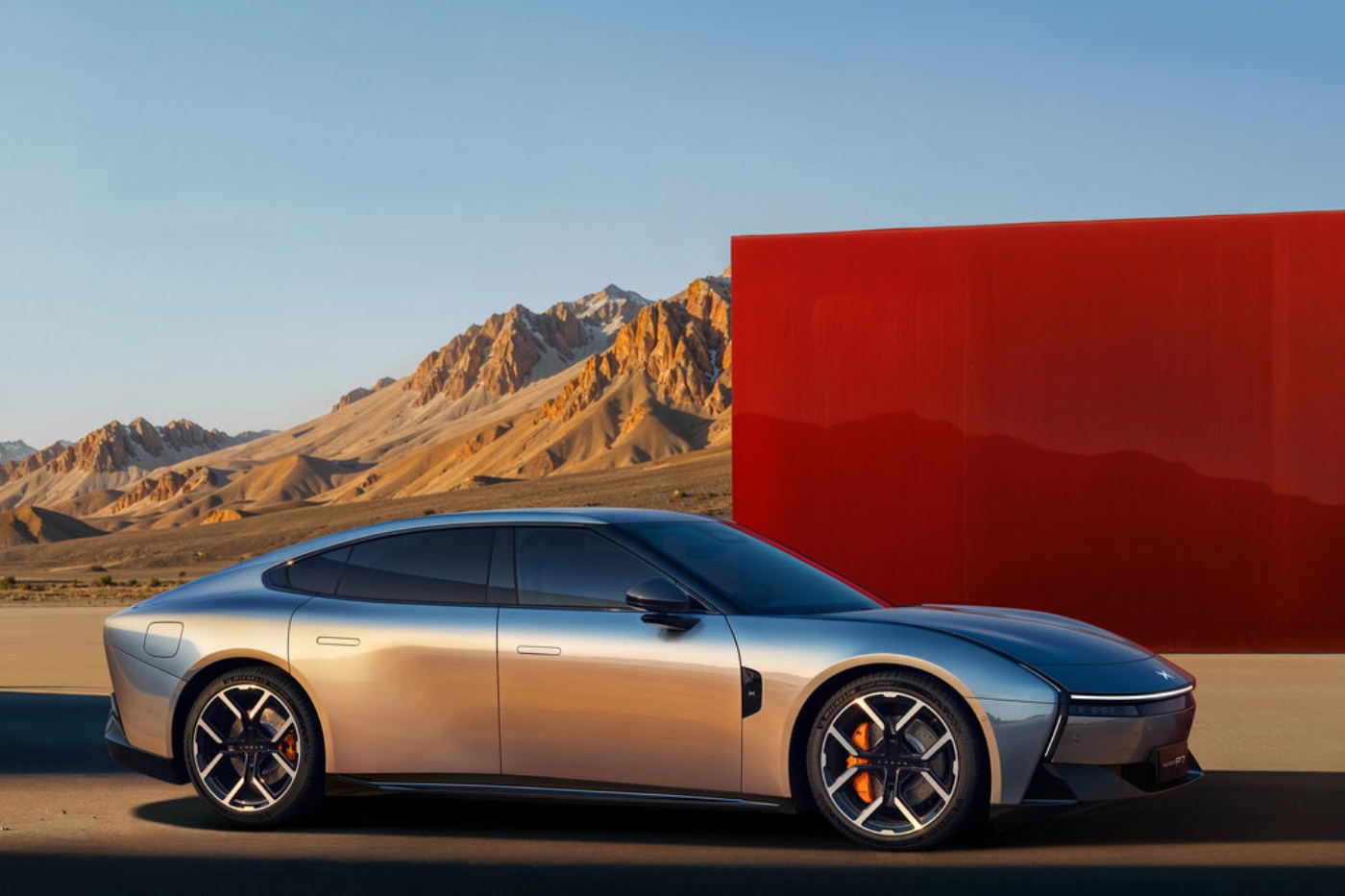 Silver electric sports sedan in a desert landscape with mountains and a red wall in the background.