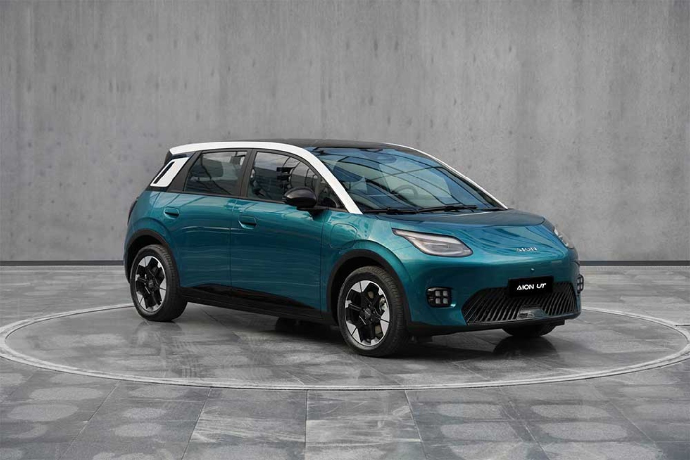 Compact electric SUV in teal color displayed on a circular platform with a gray concrete wall.