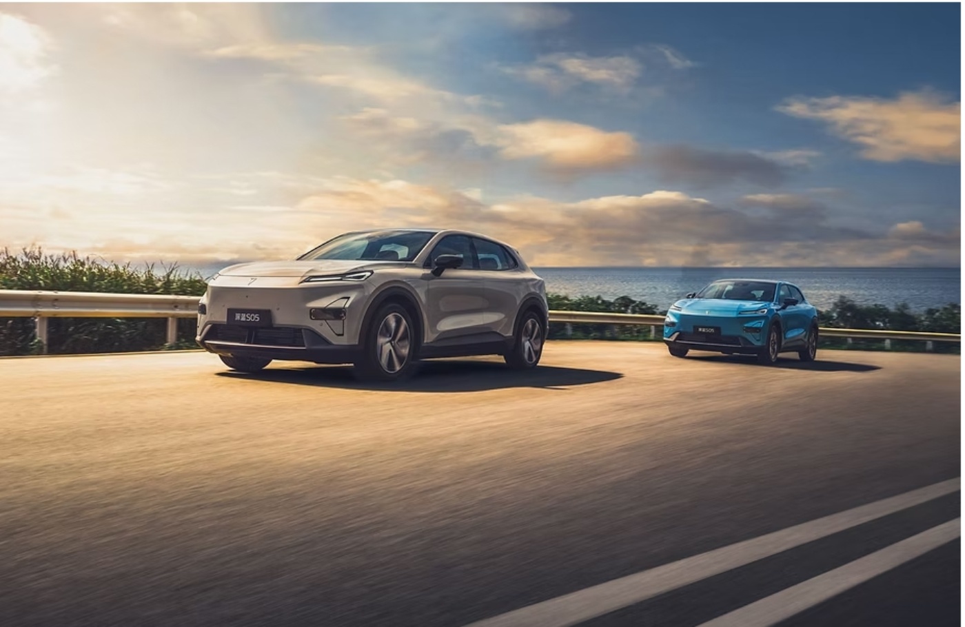 Two electric cars drive along a coastal road at sunset with ocean and clouds in the background.