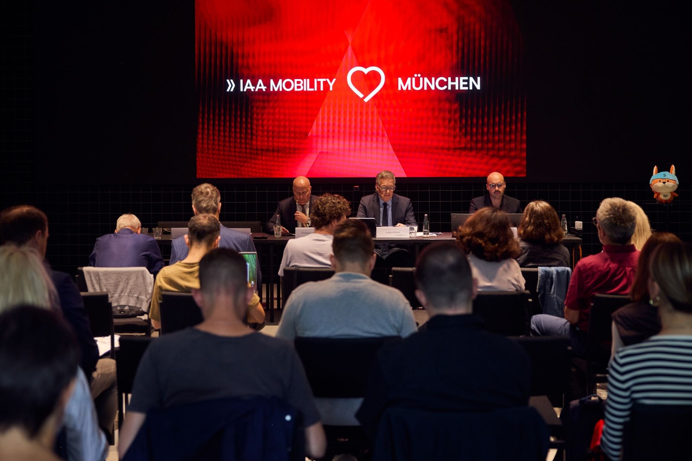 Podiumsdiskussion bei der IAA Mobility in München mit Publikum und rotem Bildschirm im Hintergrund.