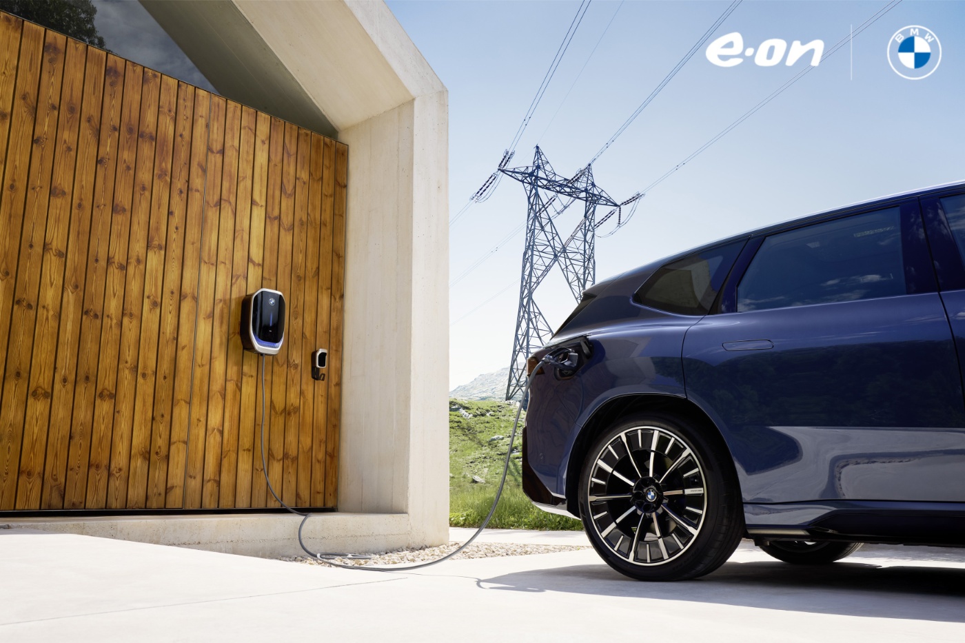 Elektrisches BMW-Fahrzeug lädt an einer Wallbox vor Holzfassade nahe Stromleitungen im Grünen.
