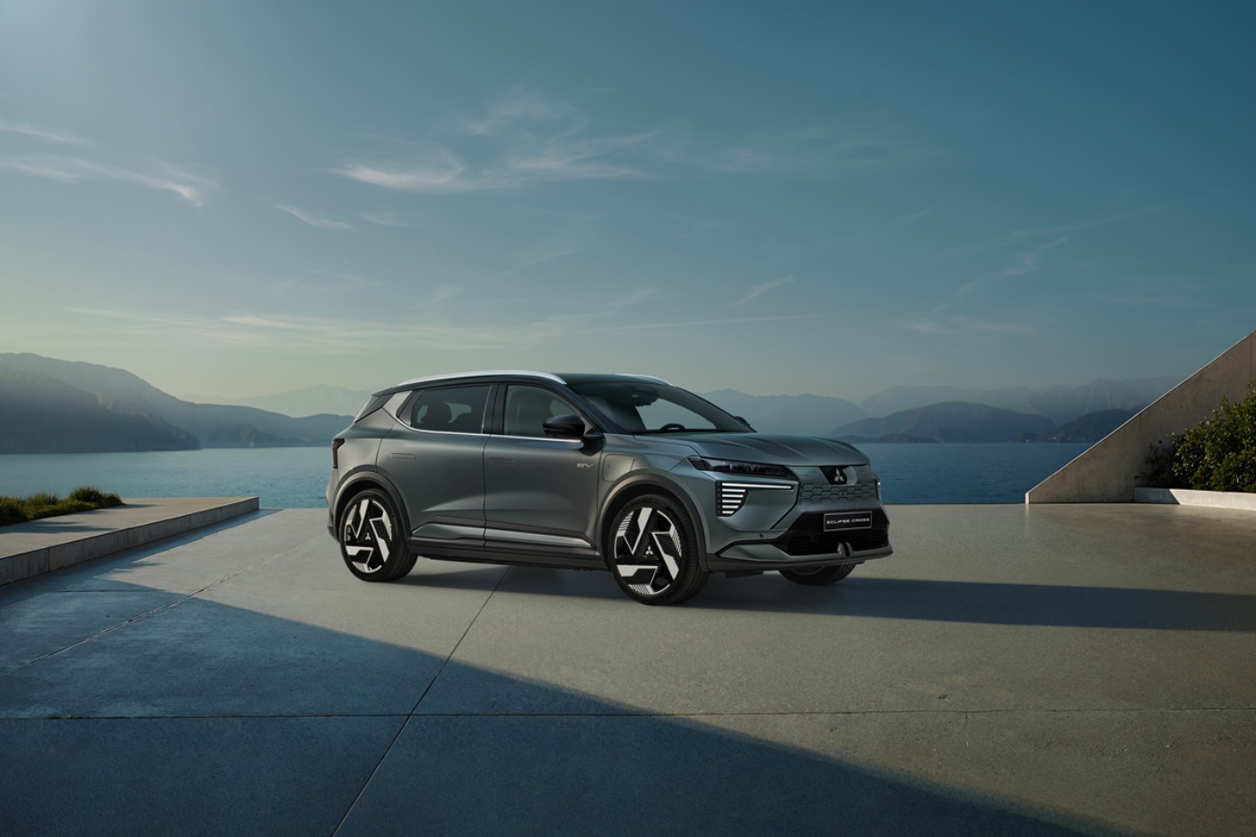 A silver Mitsubishi SUV is parked on a modern terrace with a scenic lake and mountains behind.