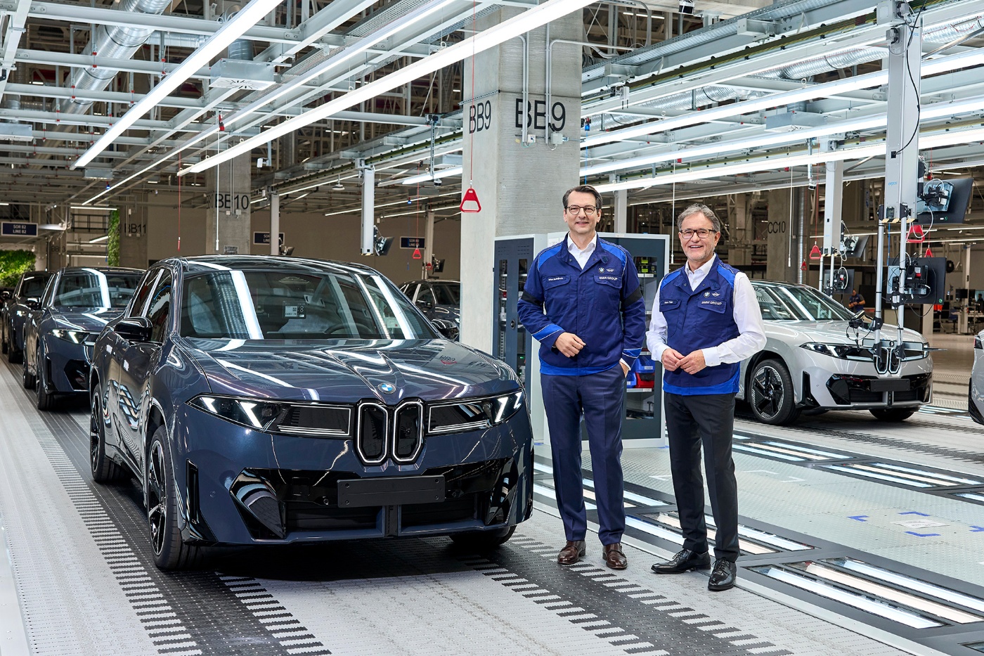Zwei Männer in BMW-Arbeitskleidung stehen vor E-Autos auf einem Montageband in einer Fabrikhalle.