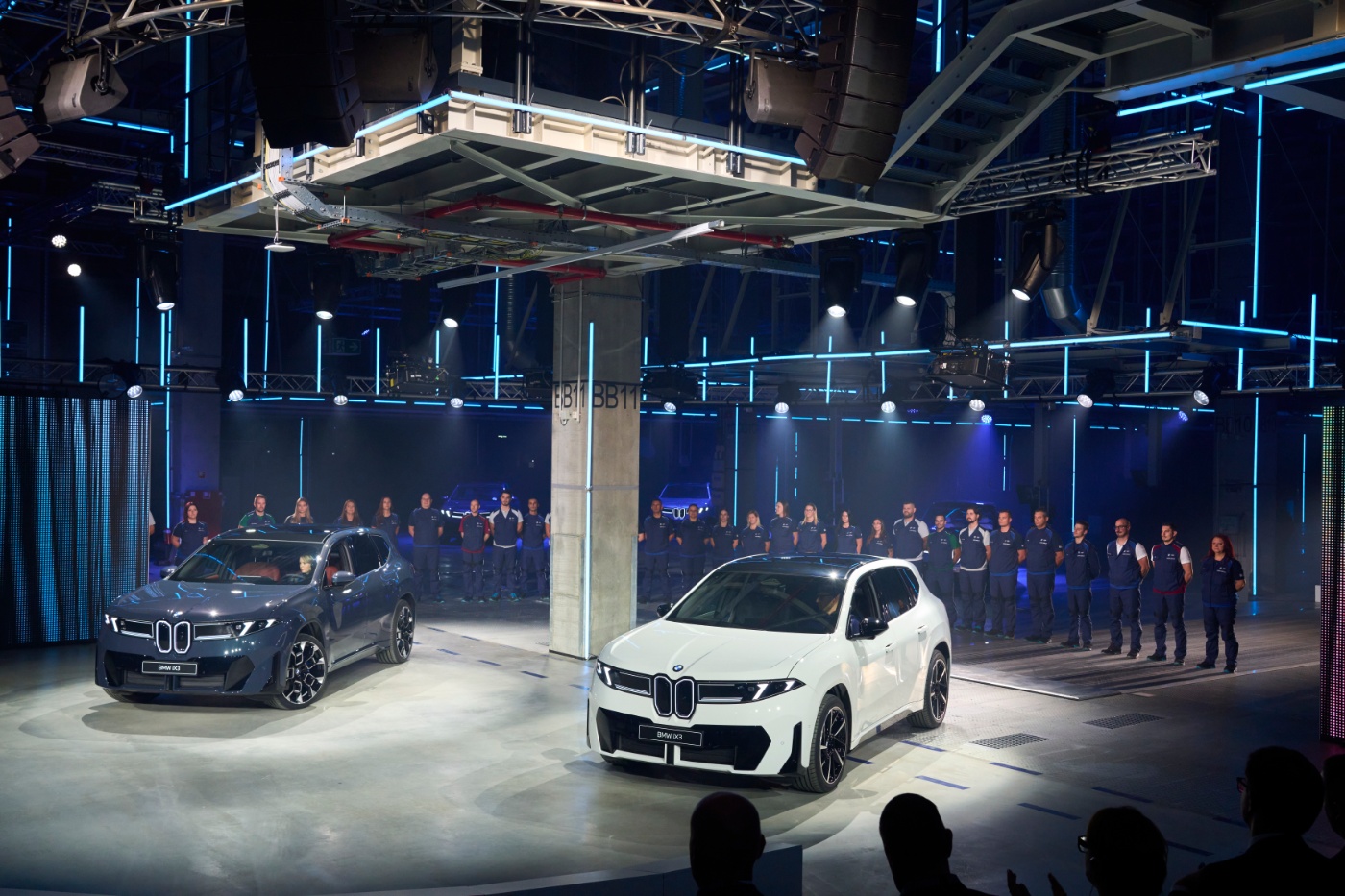 Two BMW iX3 cars are presented indoors with factory workers lined up in the background.