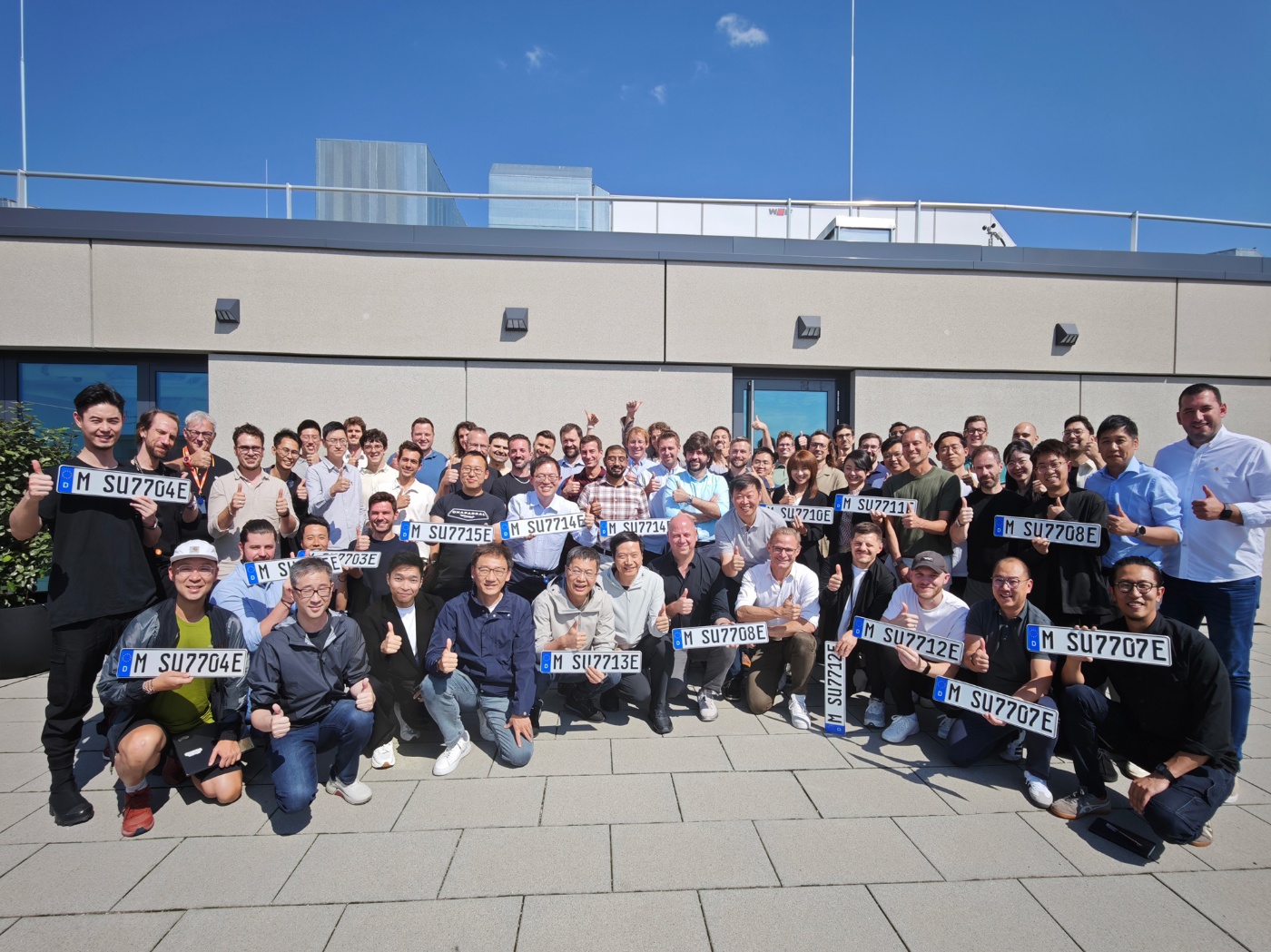 A large group poses outdoors holding license plates with thumbs up in front of a modern building.