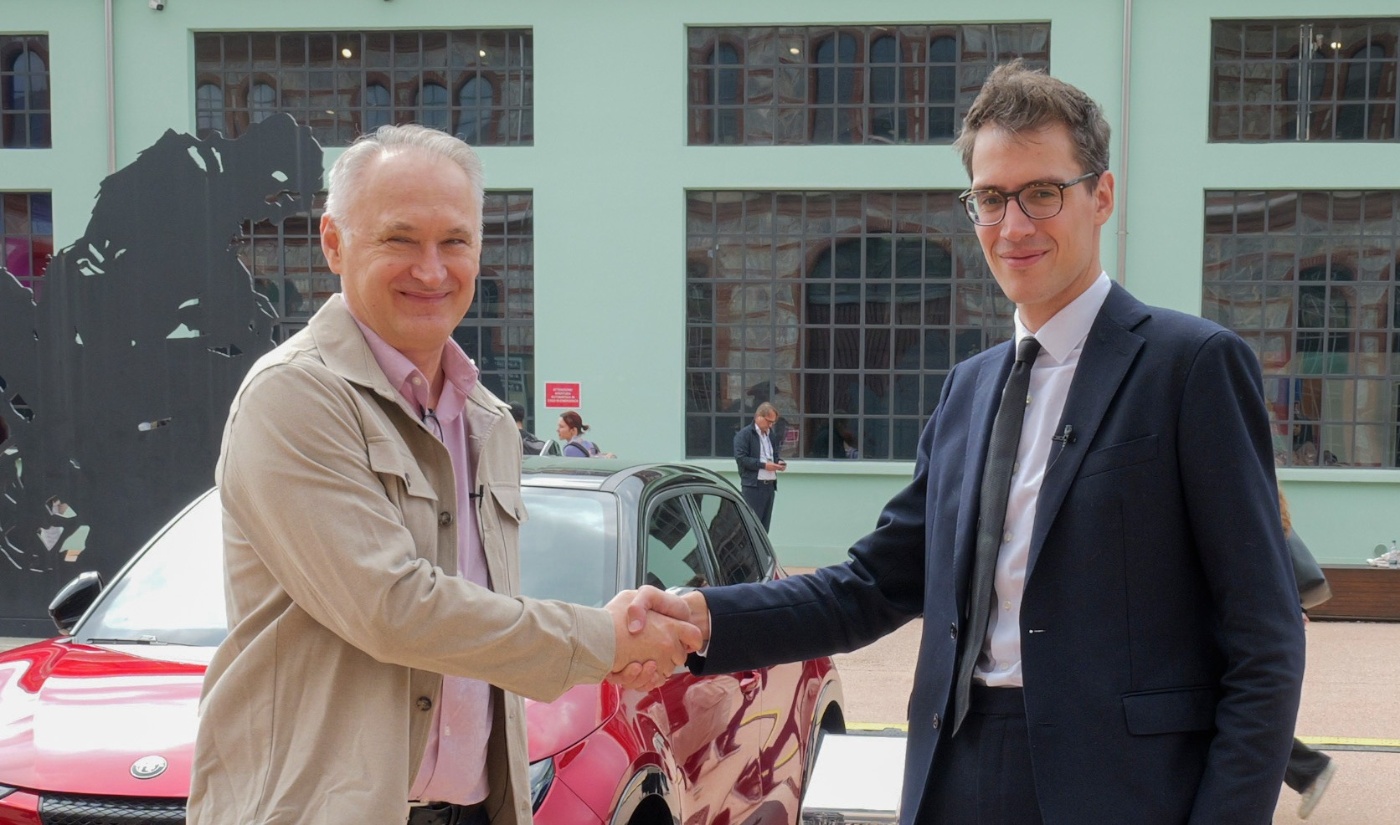 Two men shake hands in front of a red car, standing outside a building with large windows.