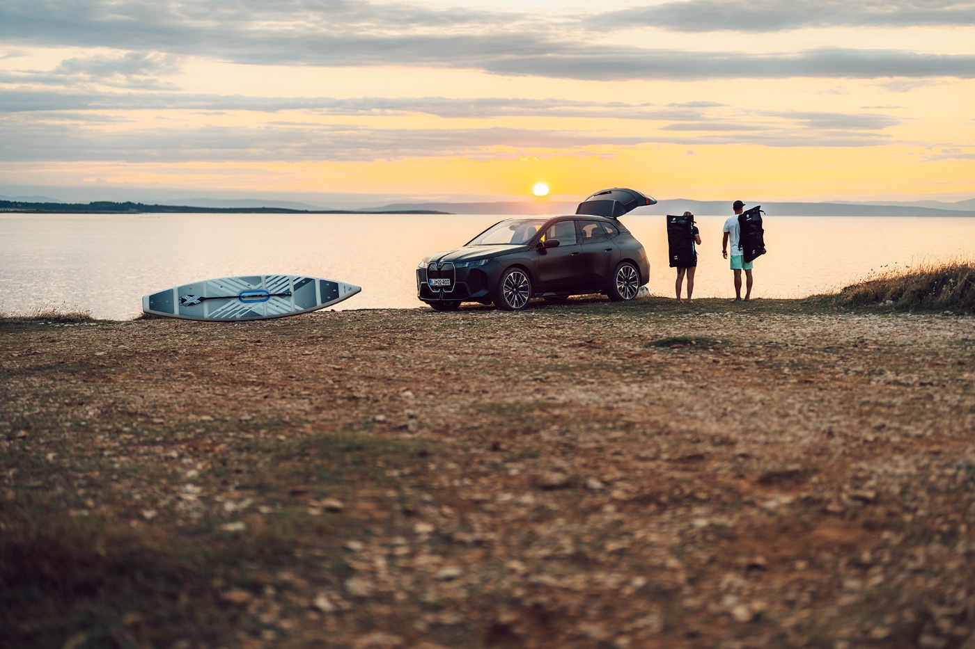 Two people with gear beside a BMW SUV and paddleboard by a lake at sunset.
