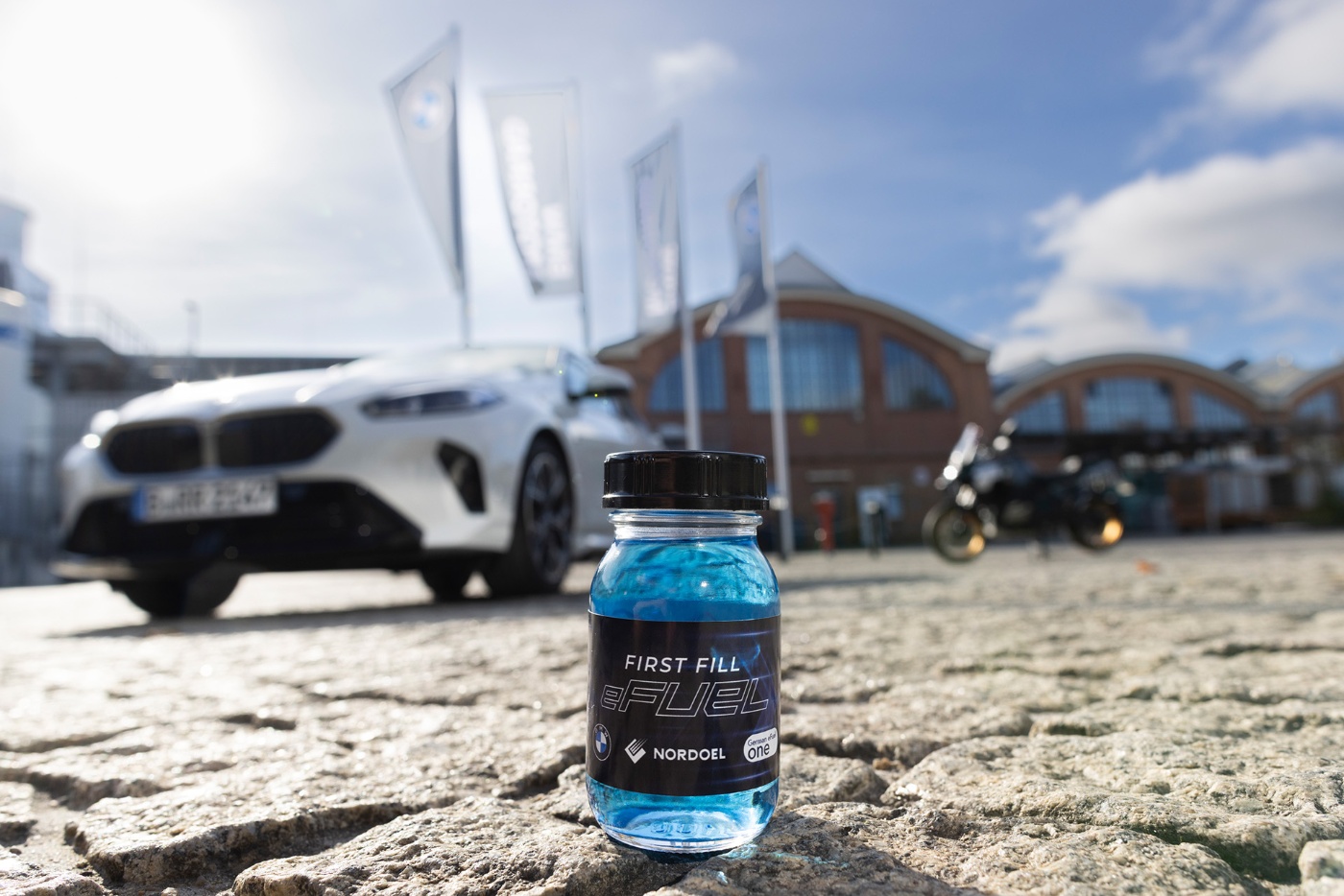 Ein kleines Fläschchen mit blauem E-Fuel steht im Vordergrund vor einem unscharfen BMW und Motorrad.
