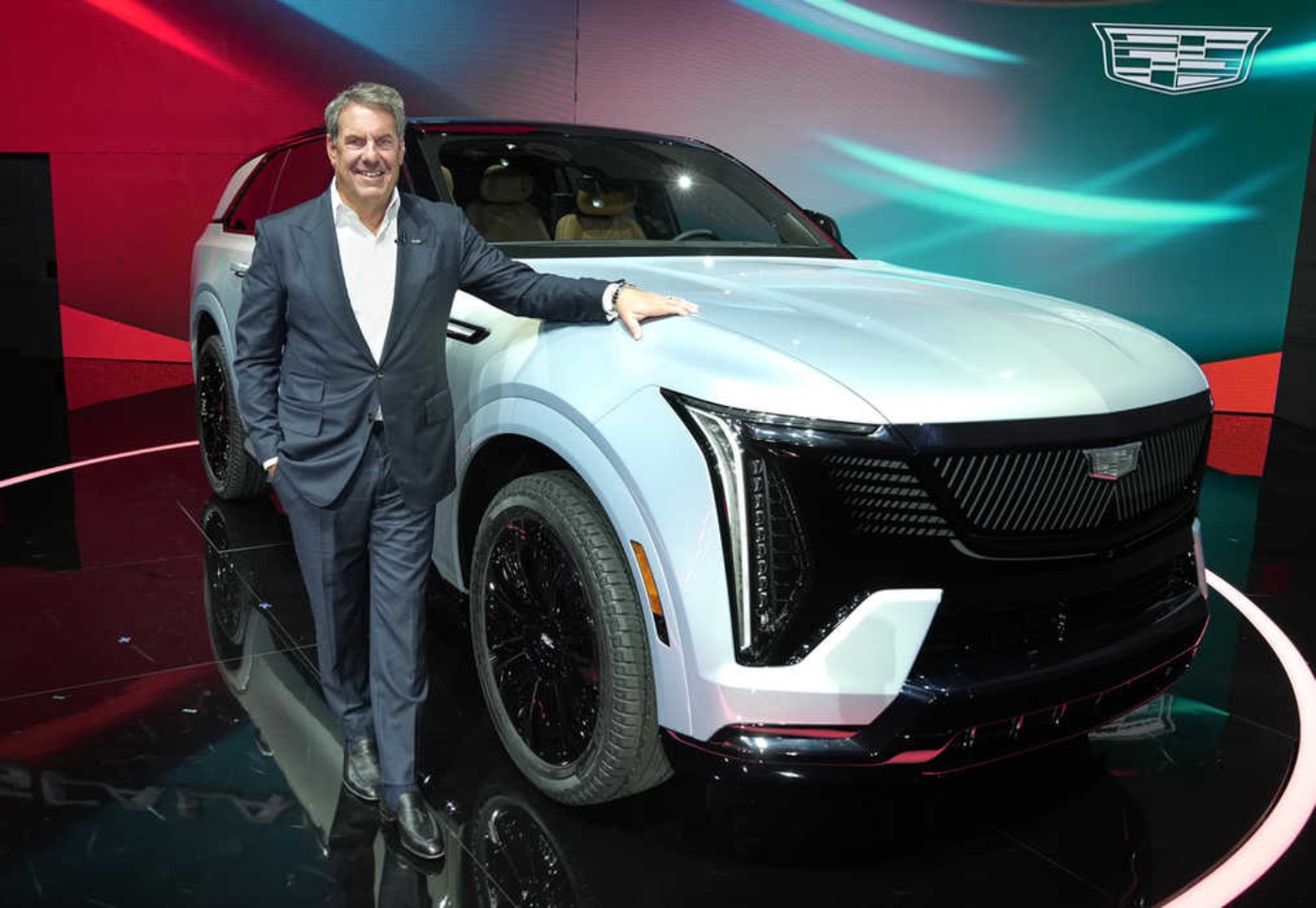 Ein Mann im Anzug präsentiert einen weißen Cadillac SUV auf einer Bühne mit Markenlogo im Hintergrund.