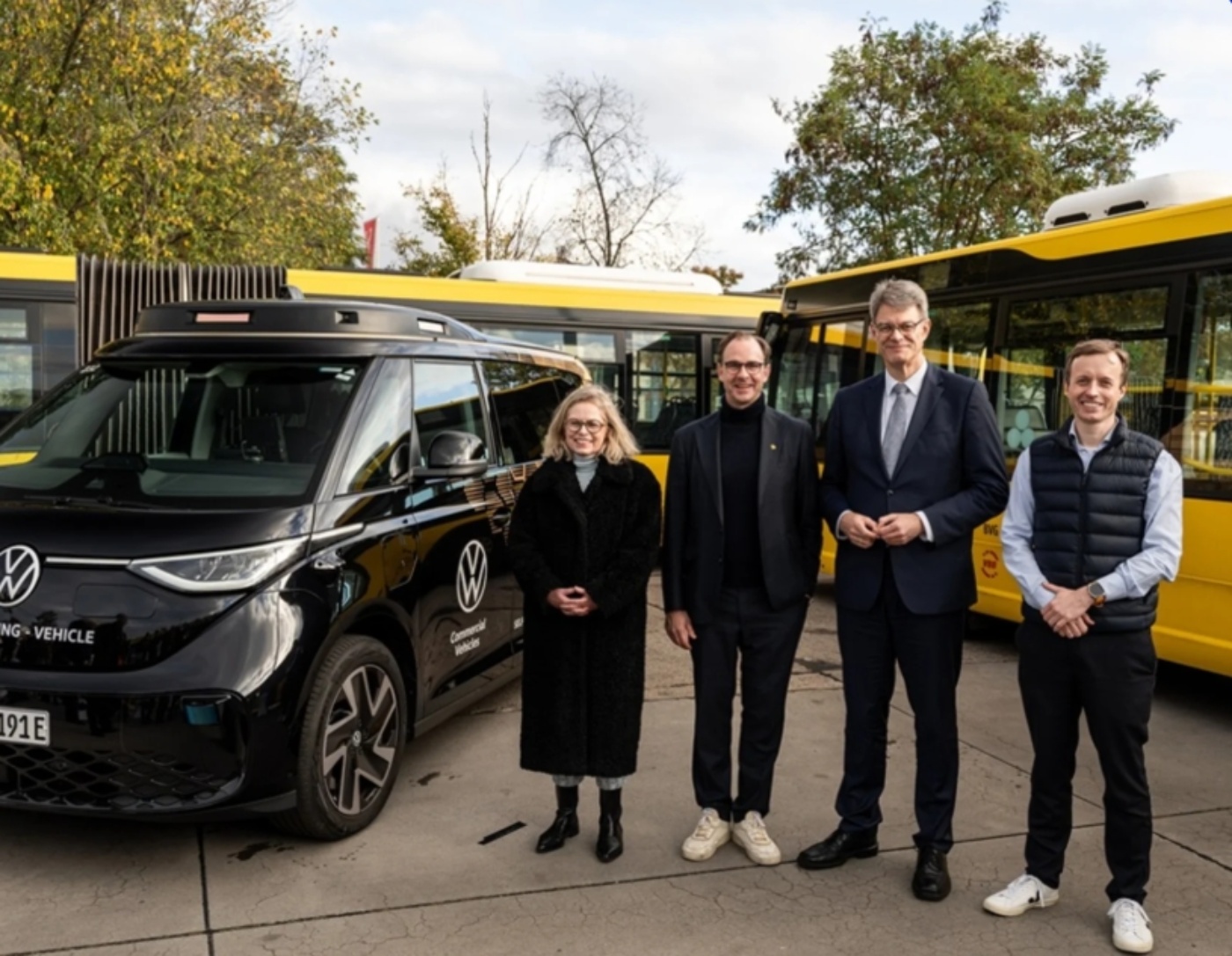 Vier Personen stehen vor einem schwarzen VW-Shuttle und gelben Bussen an einem ÖPNV-Standort.
