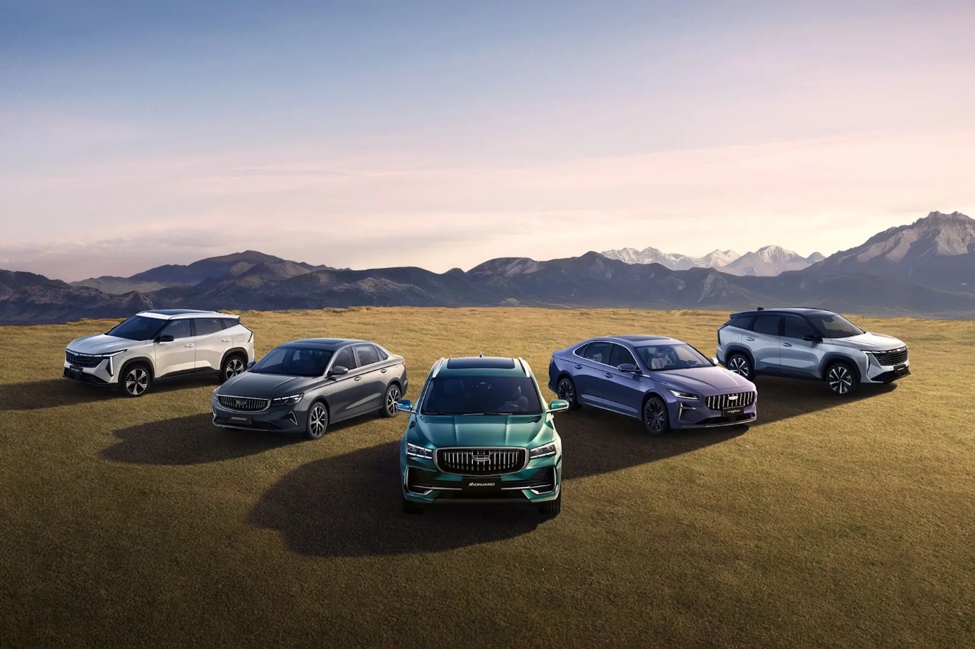 Five Geely cars of different models and colors are parked on a grassy field with mountains in the background under a cloudy sky.